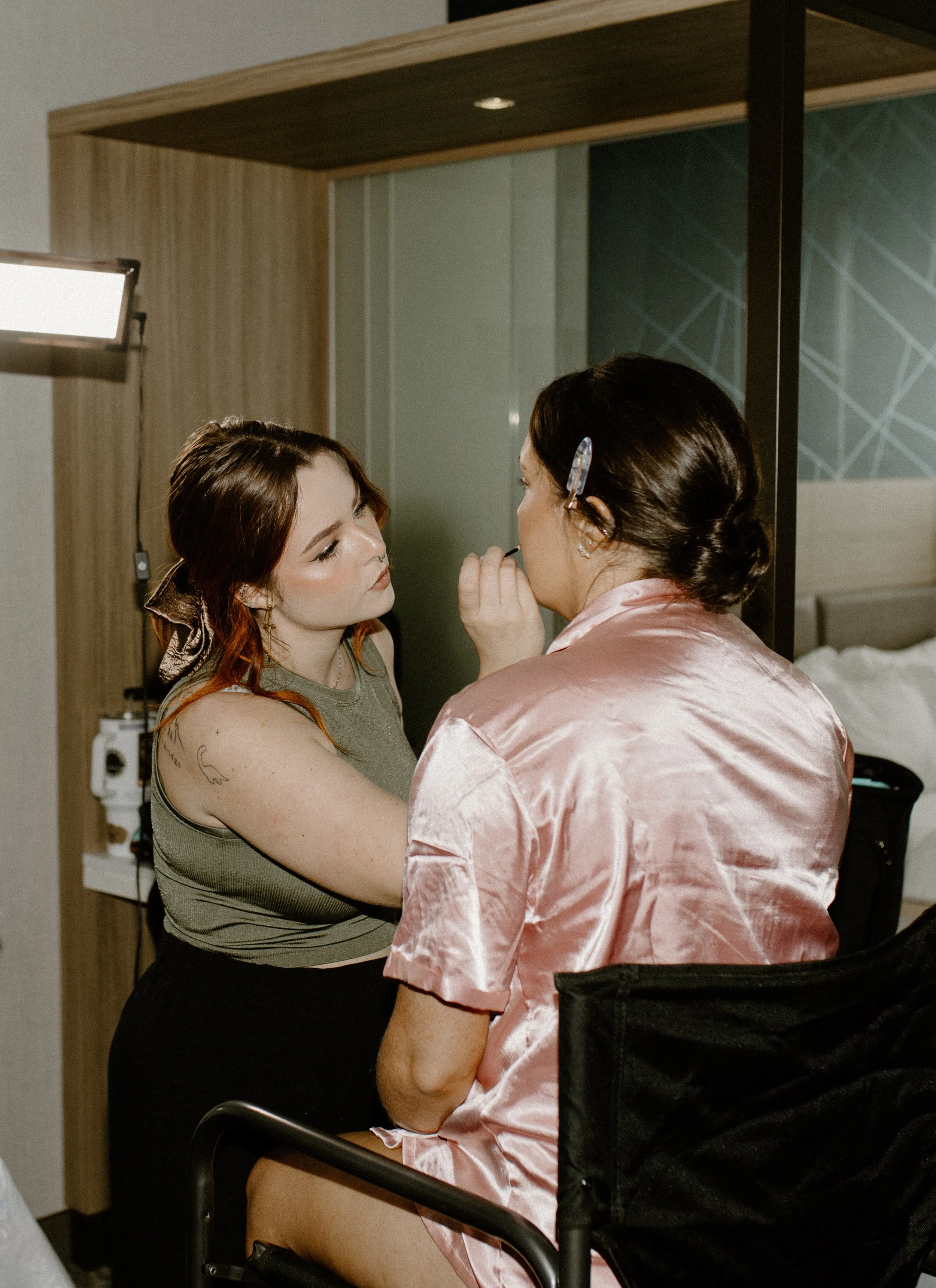 Two women in a hotel room, one applying makeup to the other.