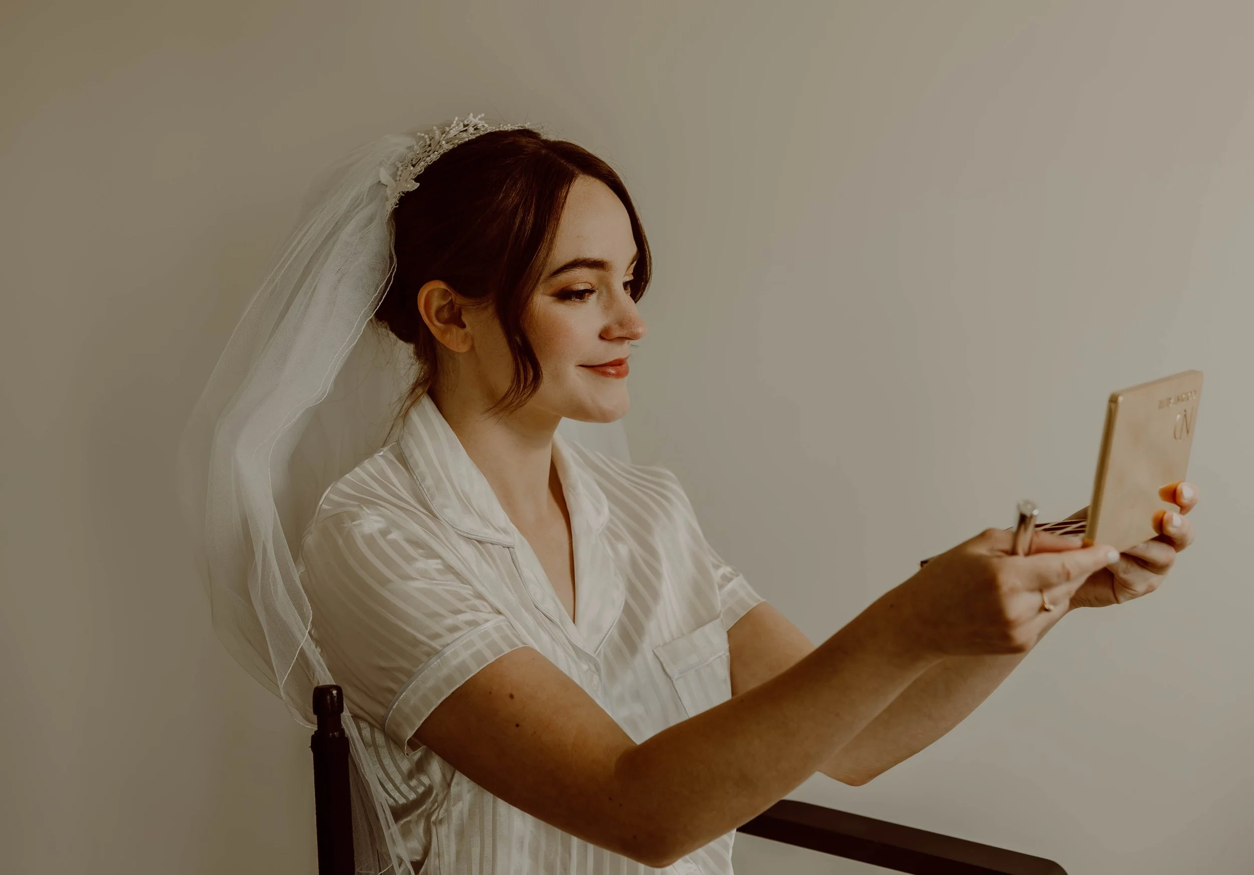 A woman with red hair, wearing a white satin robe and a wedding veil, sitting and looking at her phone.