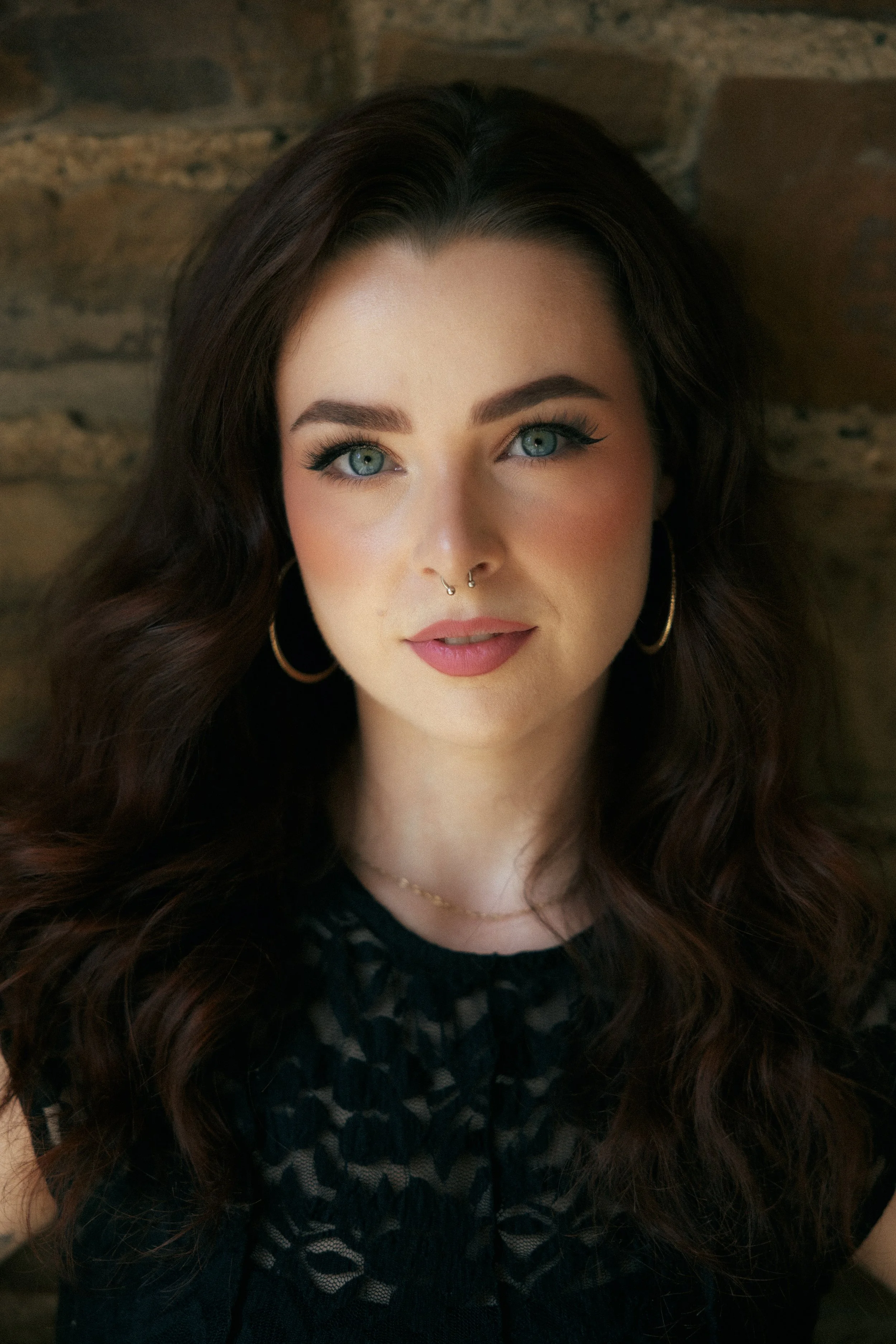 A young woman with long, wavy brown hair and blue eyes, wearing gold hoop earrings and a black lace top, standing against a brick wall.