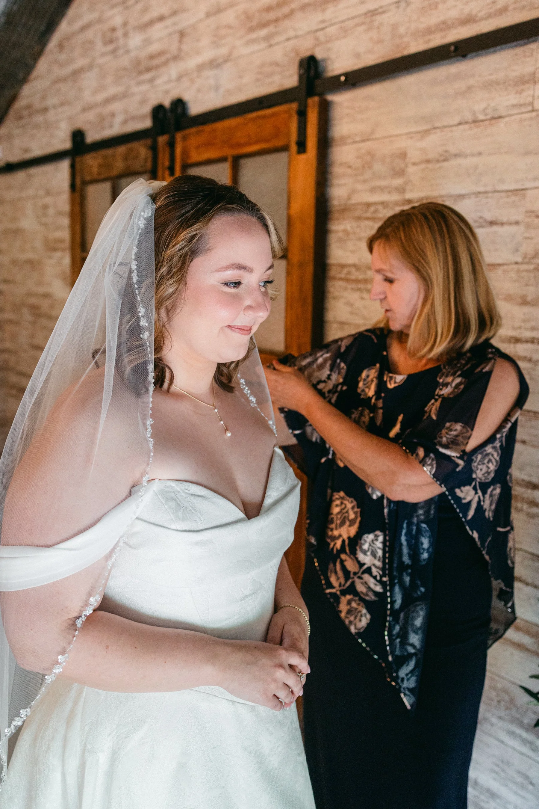A bride wearing a white wedding dress and veil, with a woman in a black floral dress helping her with her dress, in a room with wood-paneled walls.