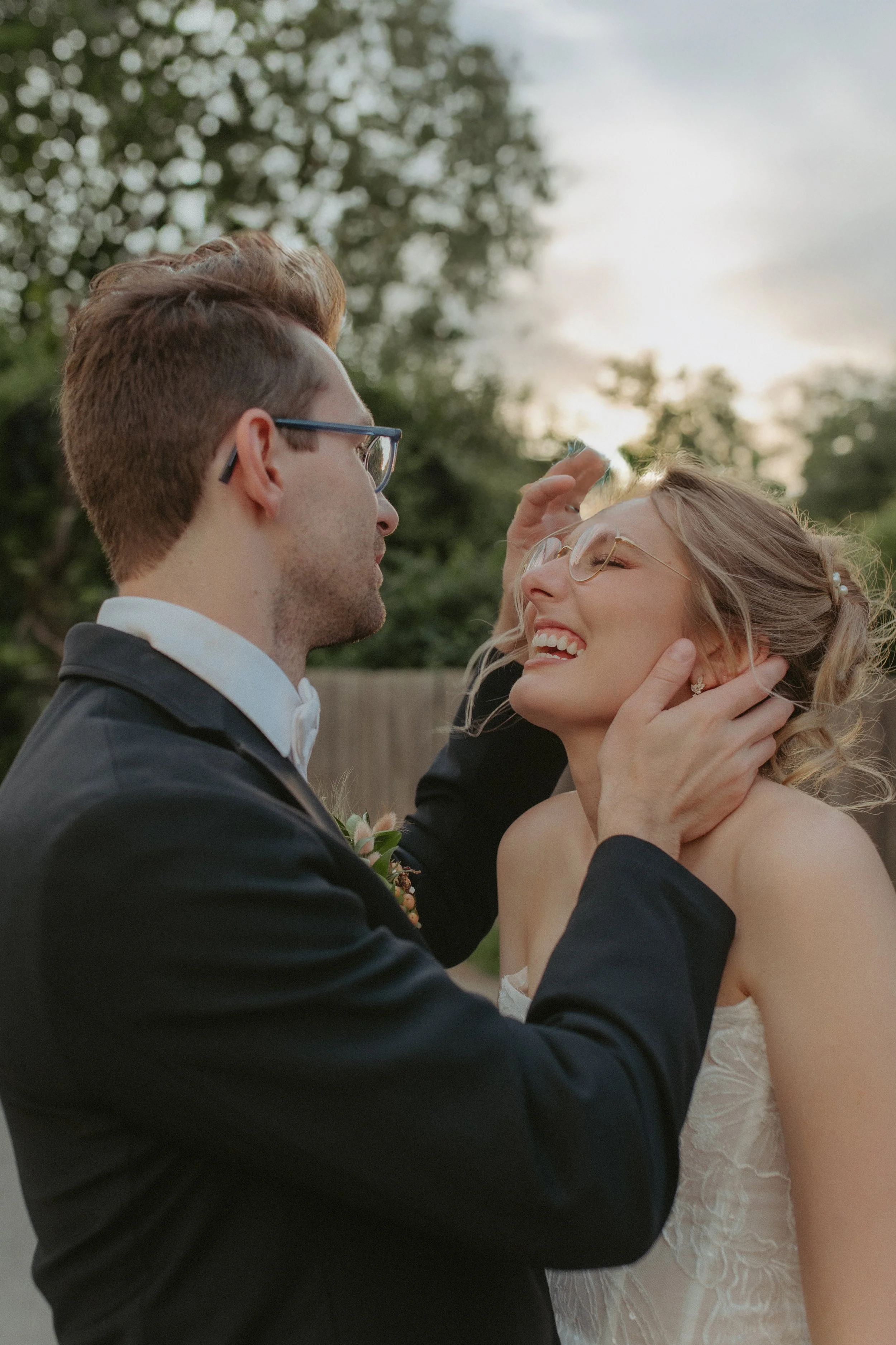 A couple dressed in wedding attire sharing a moment outdoors with trees in the background, during sunset.