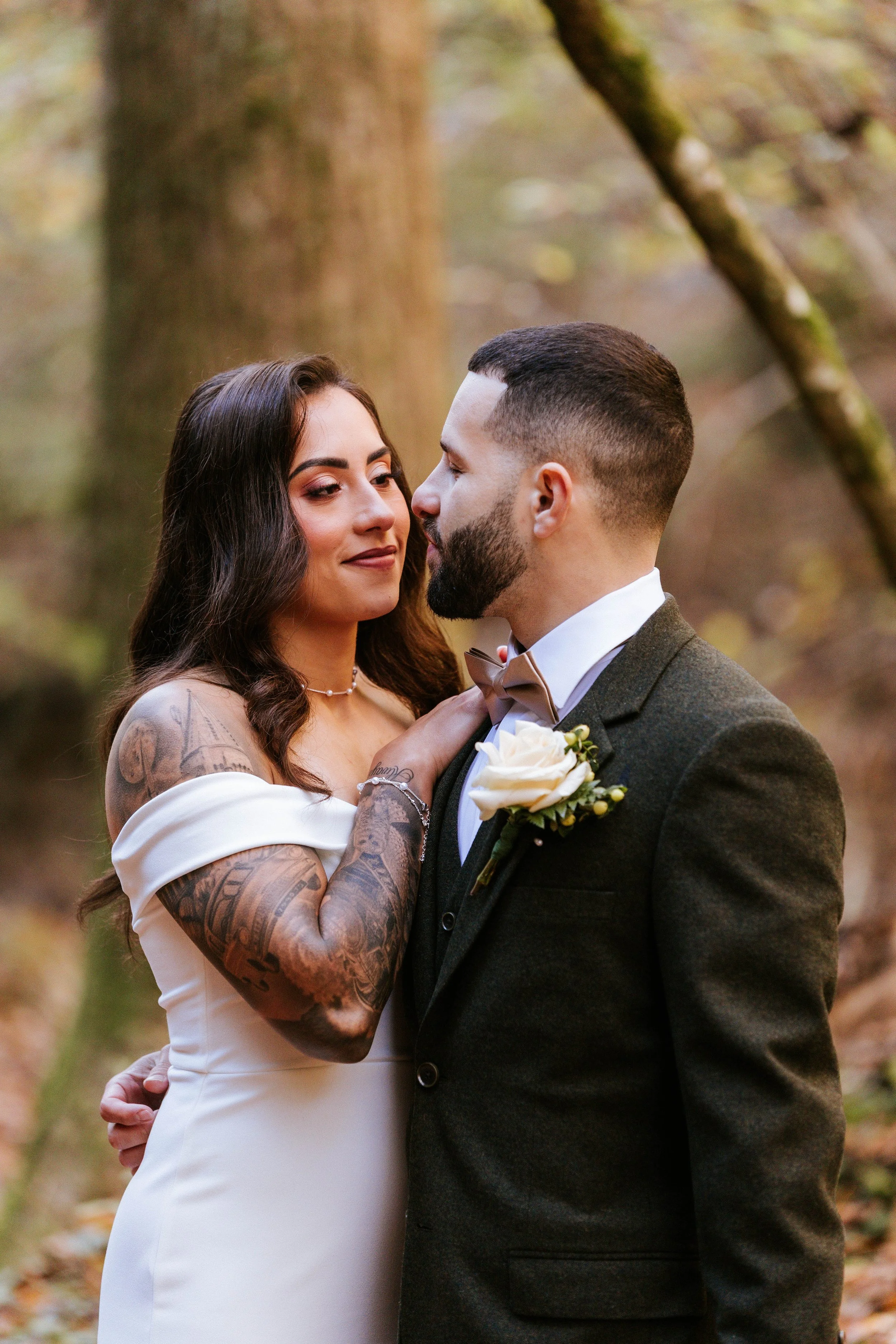 A couple dressed in wedding attire sharing an intimate moment outdoors, with autumn trees and fallen leaves in the background.