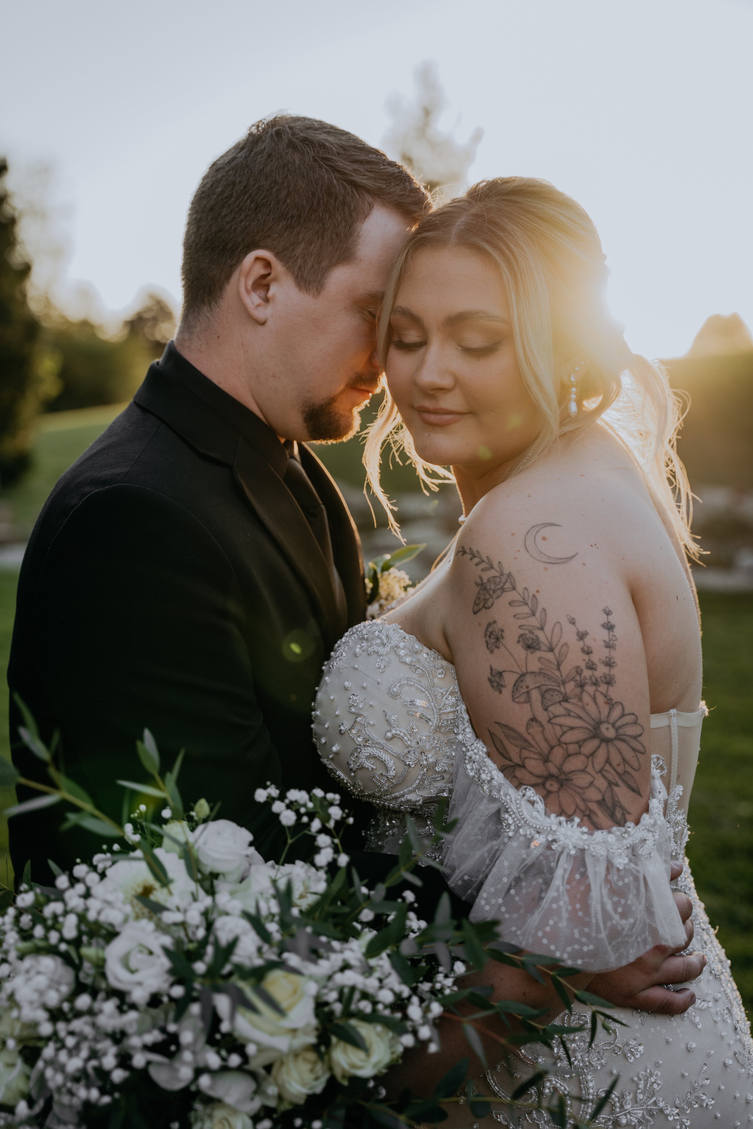 A bride and groom share an intimate moment outdoors during sunset, with the bride having tattoos on her arm and wearing a white lace gown, and the groom in a black suit. The bride holds a bouquet of white flowers and greenery.