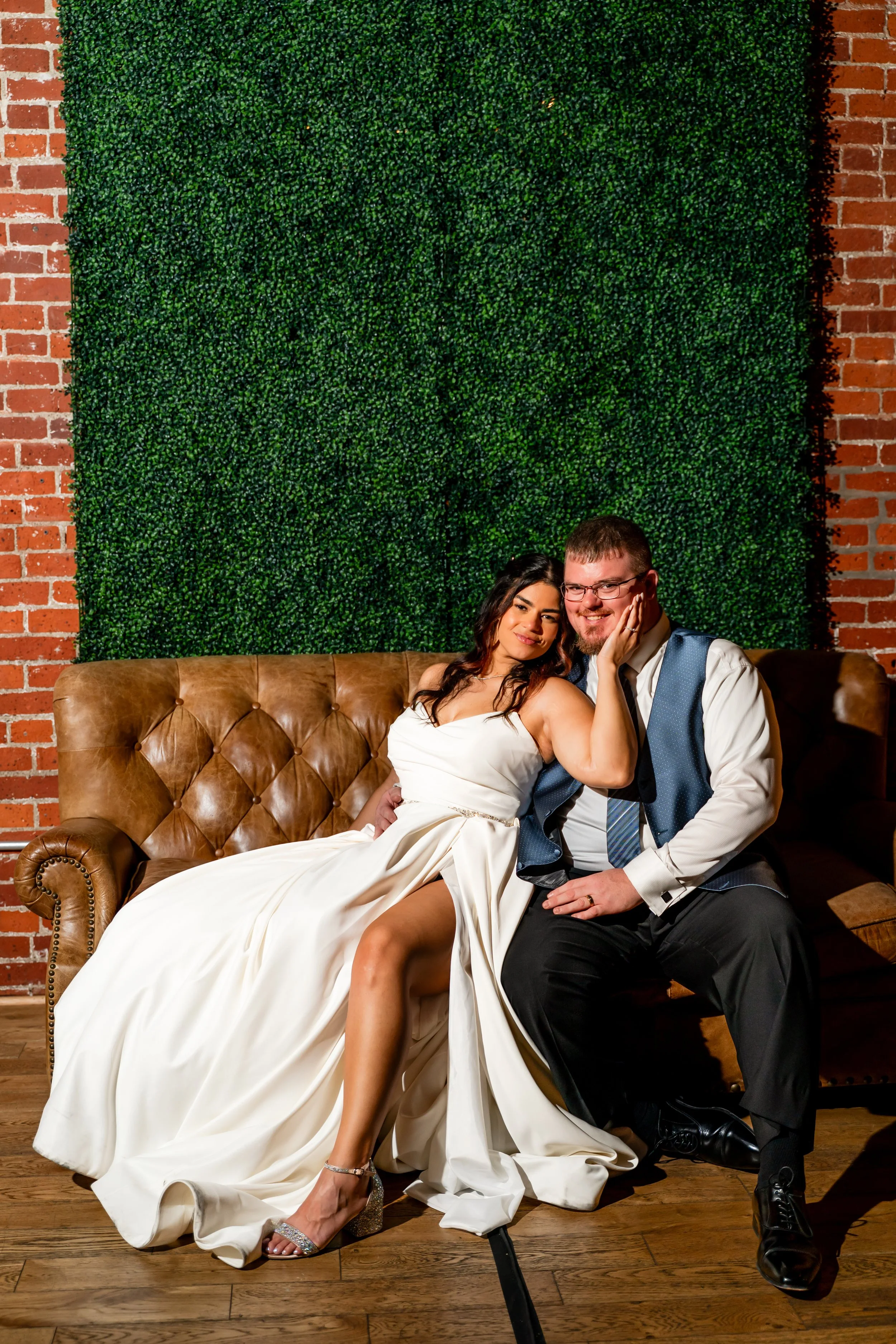 A couple dressed in wedding attire sitting on a vintage brown leather sofa. The woman is wearing a white wedding dress with a slit, silver high heels, and has dark hair. The man is wearing a dark vest, light shirt, striped tie, glasses, and has light