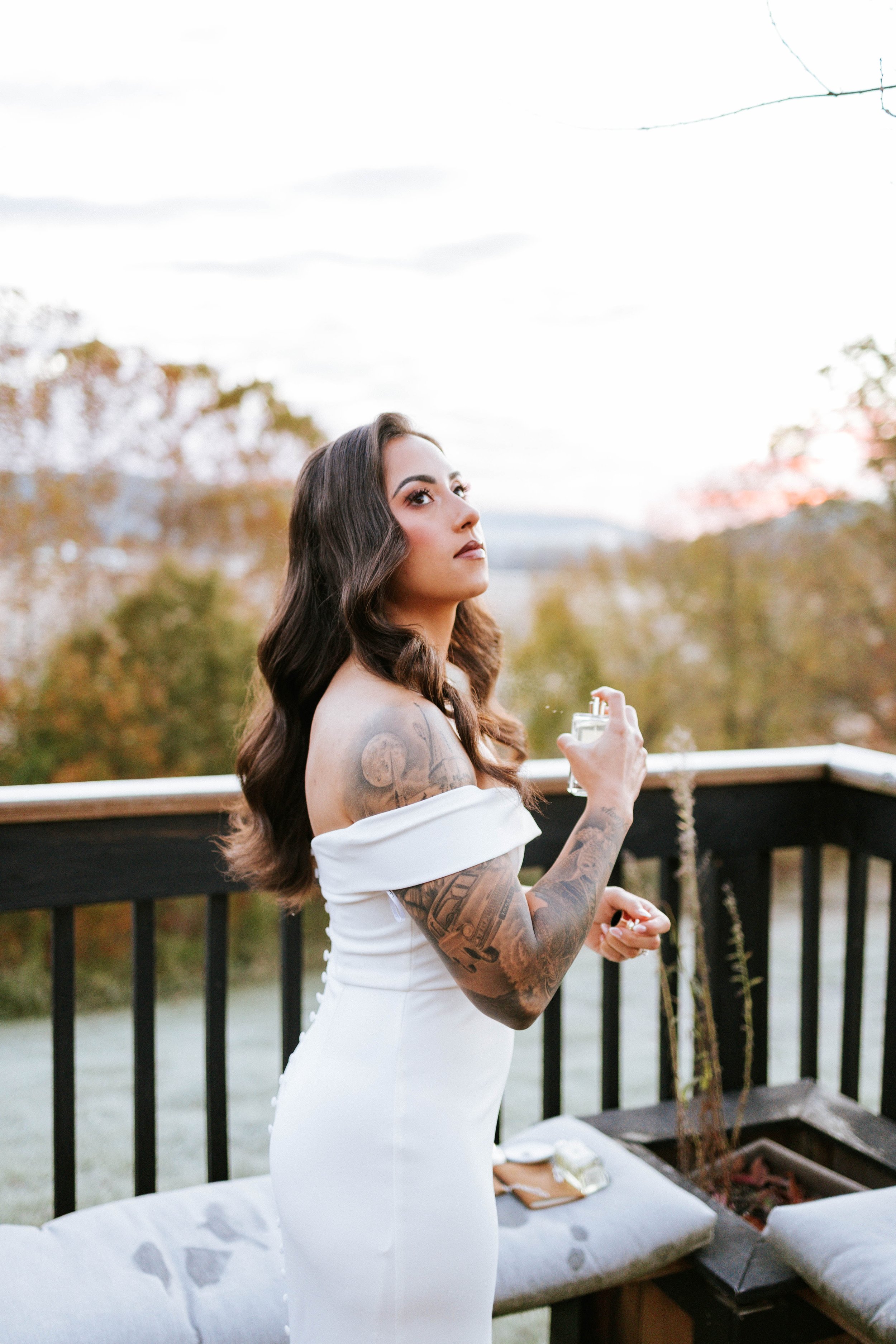 A woman in an off-the-shoulder white dress standing outdoors on a balcony with trees and a cloudy sky in the background, holding a perfume bottle.
