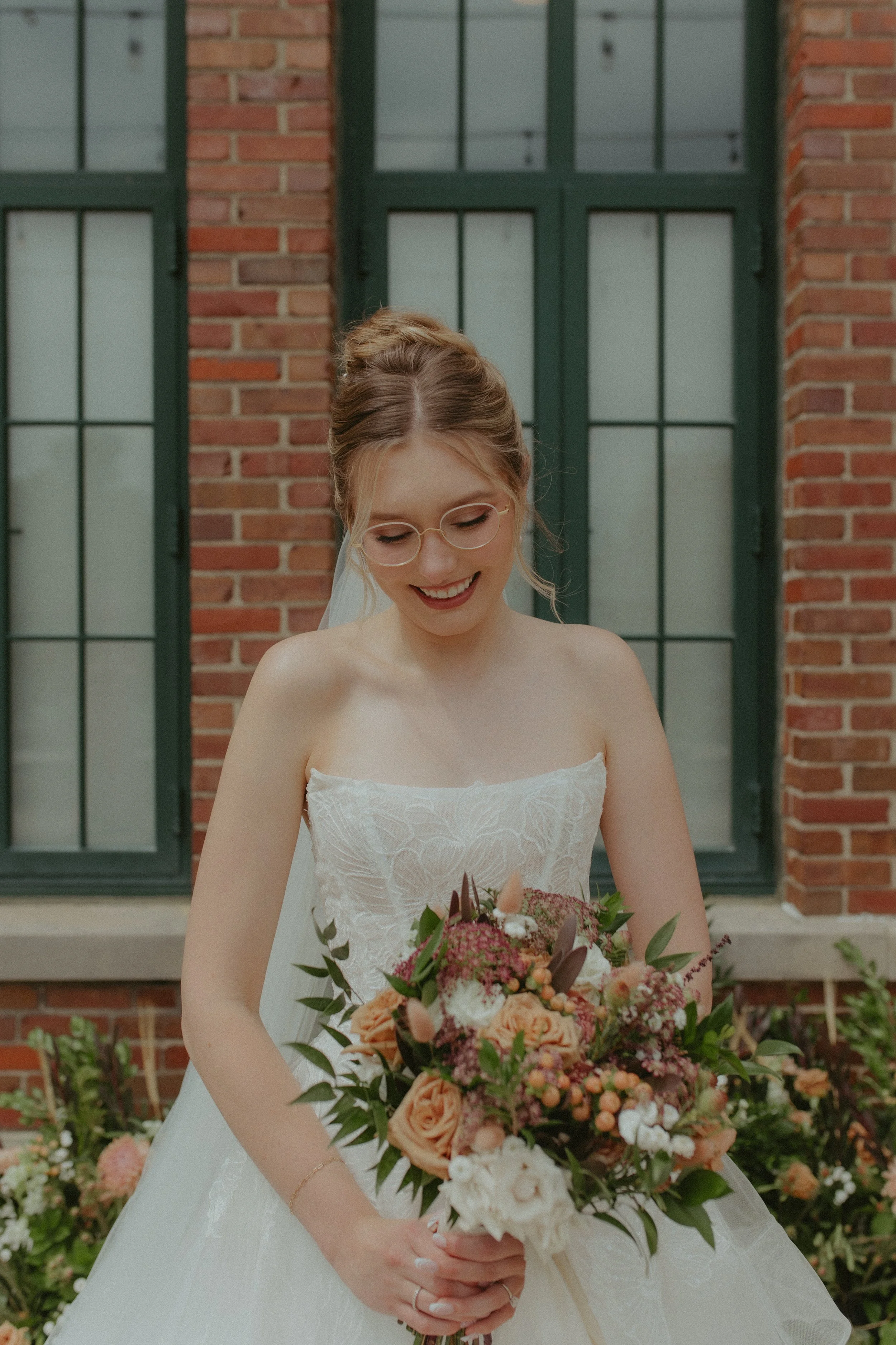 A smiling bride with blonde hair and glasses holding a large bouquet of flowers, standing in front of a brick building with large windows.