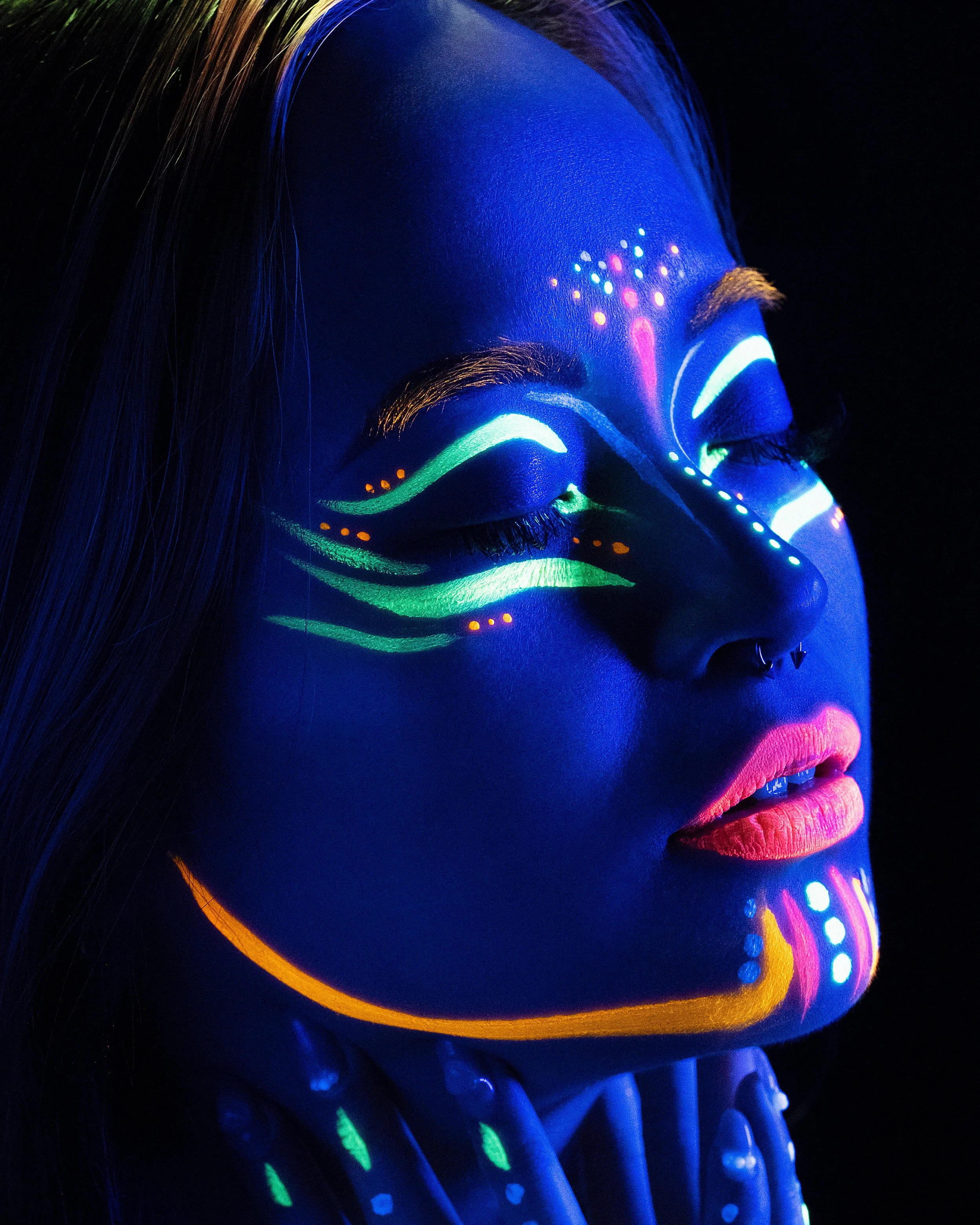 Close-up of a woman's face painted with vibrant, glowing neon colors under blacklight, emphasizing detailed patterns on her forehead, eyelids, cheeks, and lips, with long illuminated hair visible on the left side.