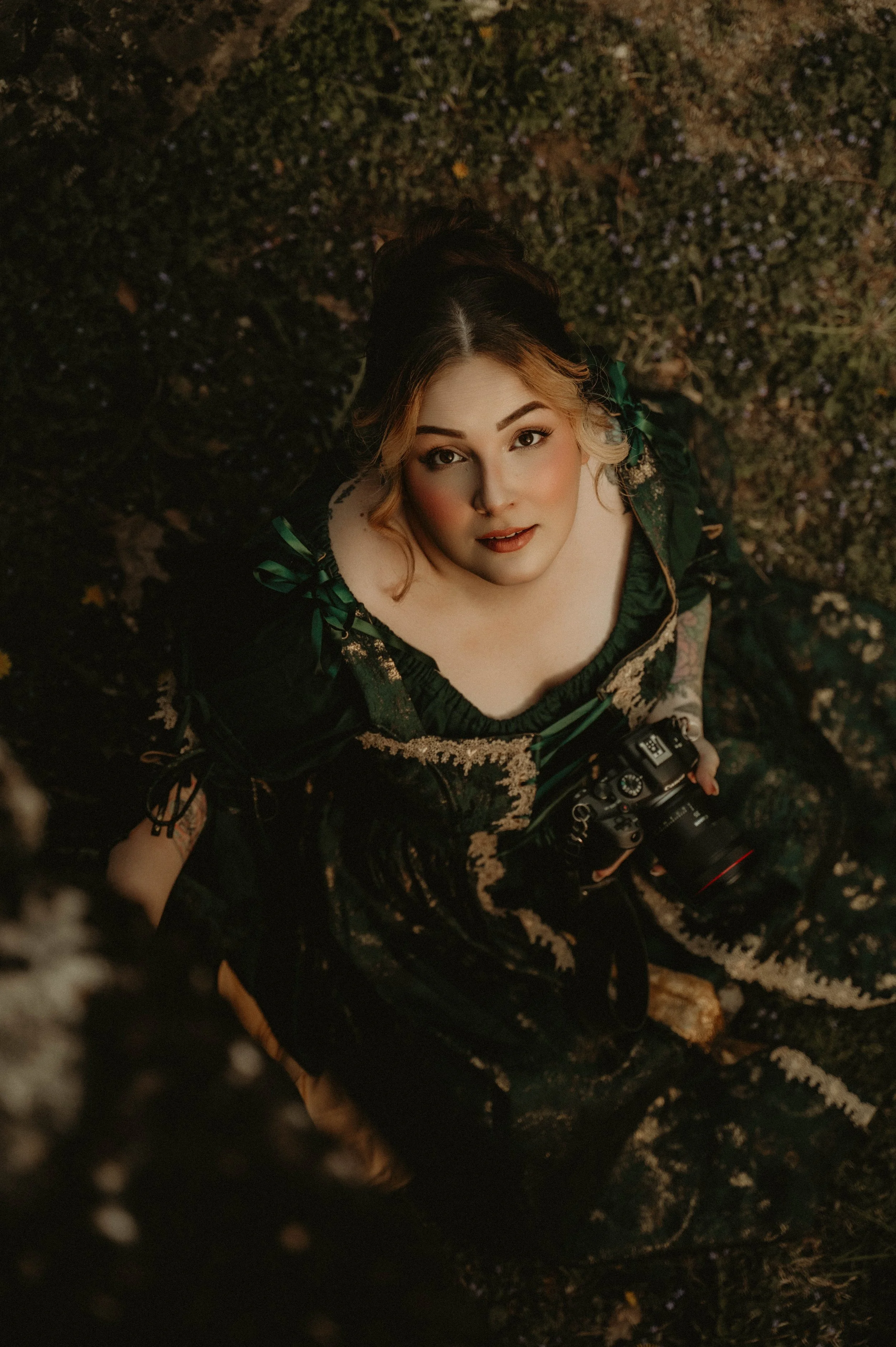 A young woman with light skin, blonde hair and makeup, looking up at the camera. She is wearing a dark green and black dress with lace details and is holding a camera in her hand. The background appears to be an outdoor setting with dark, earthy grou