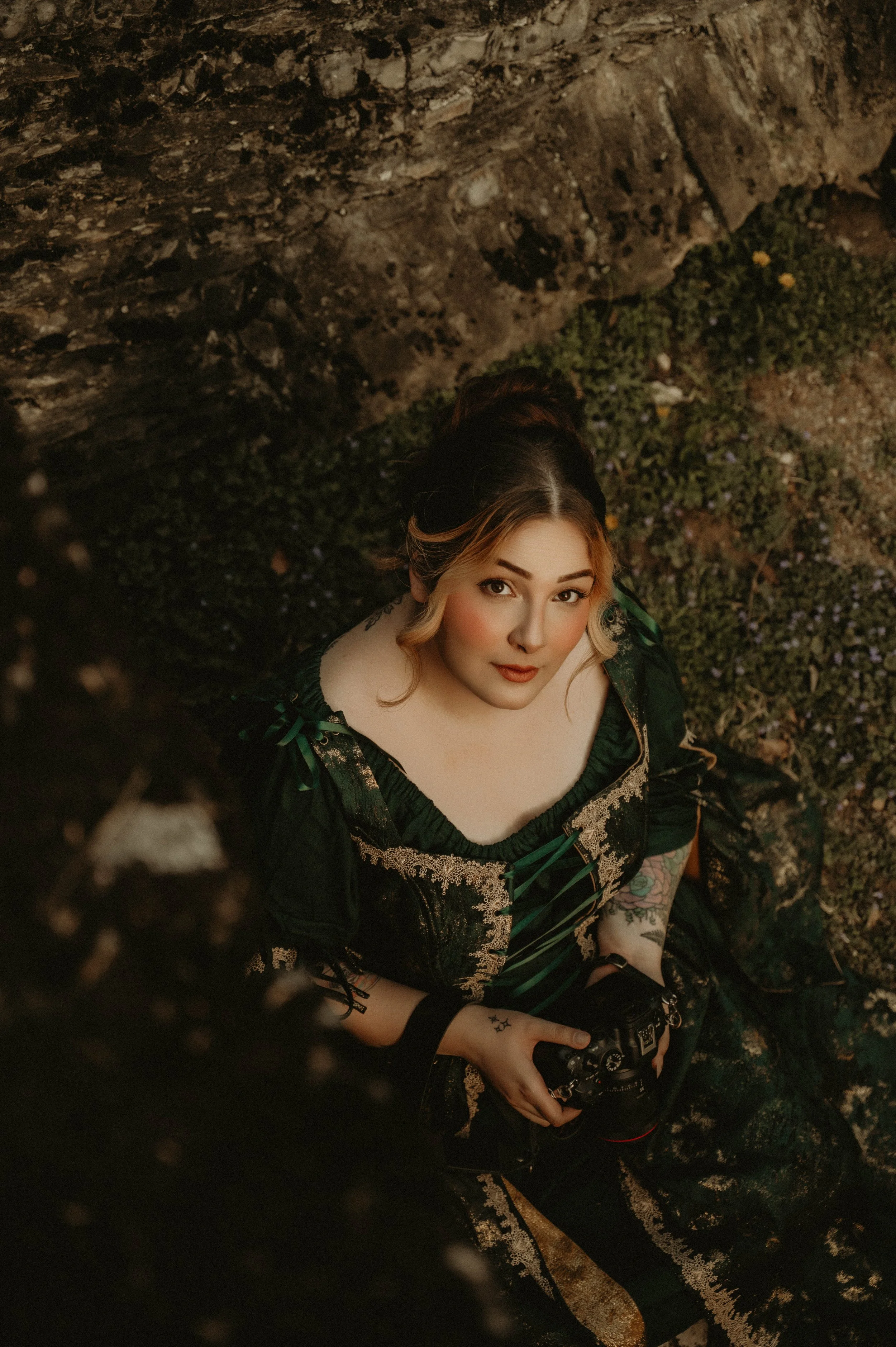 A woman with light skin, tattoos on her arm, and blonde hair with dark roots, wearing a dark green and gold dress, looking up at the camera while holding a camera in her hand.
