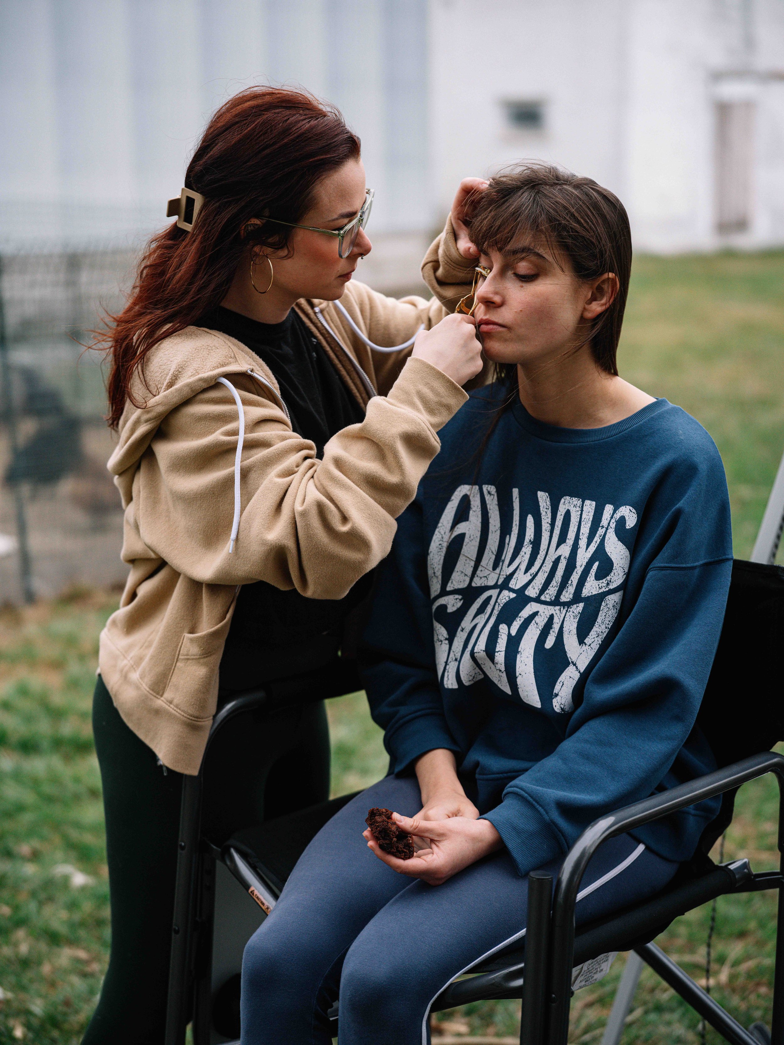 A healthcare worker in beige hoodie and glasses gives a shot to a young woman with short brown hair in a blue sweatshirt, seated in a wheelchair outdoors.