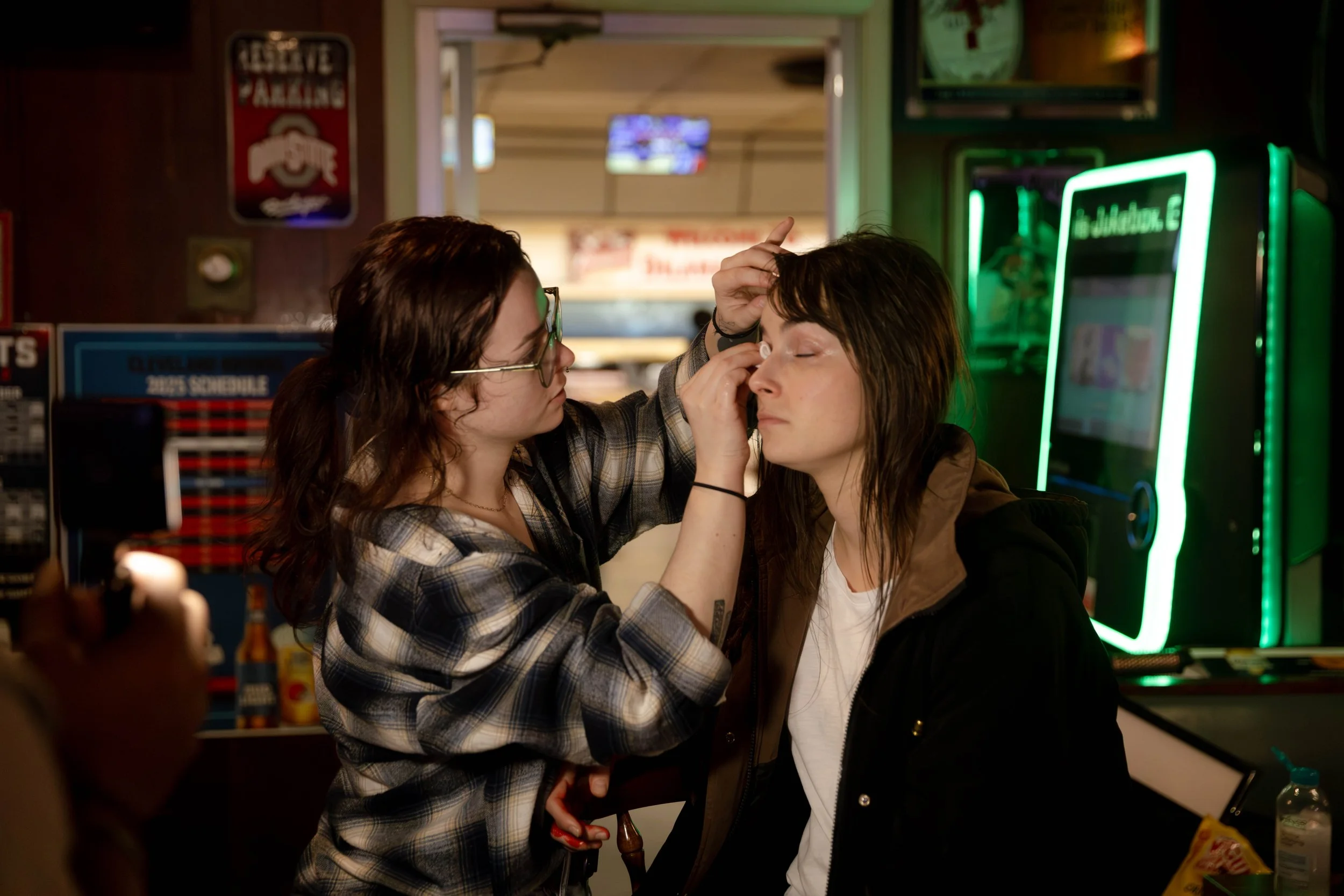 Two women in a bar with neon lights, one is applying makeup to the other's eye while the other has her eyes closed.