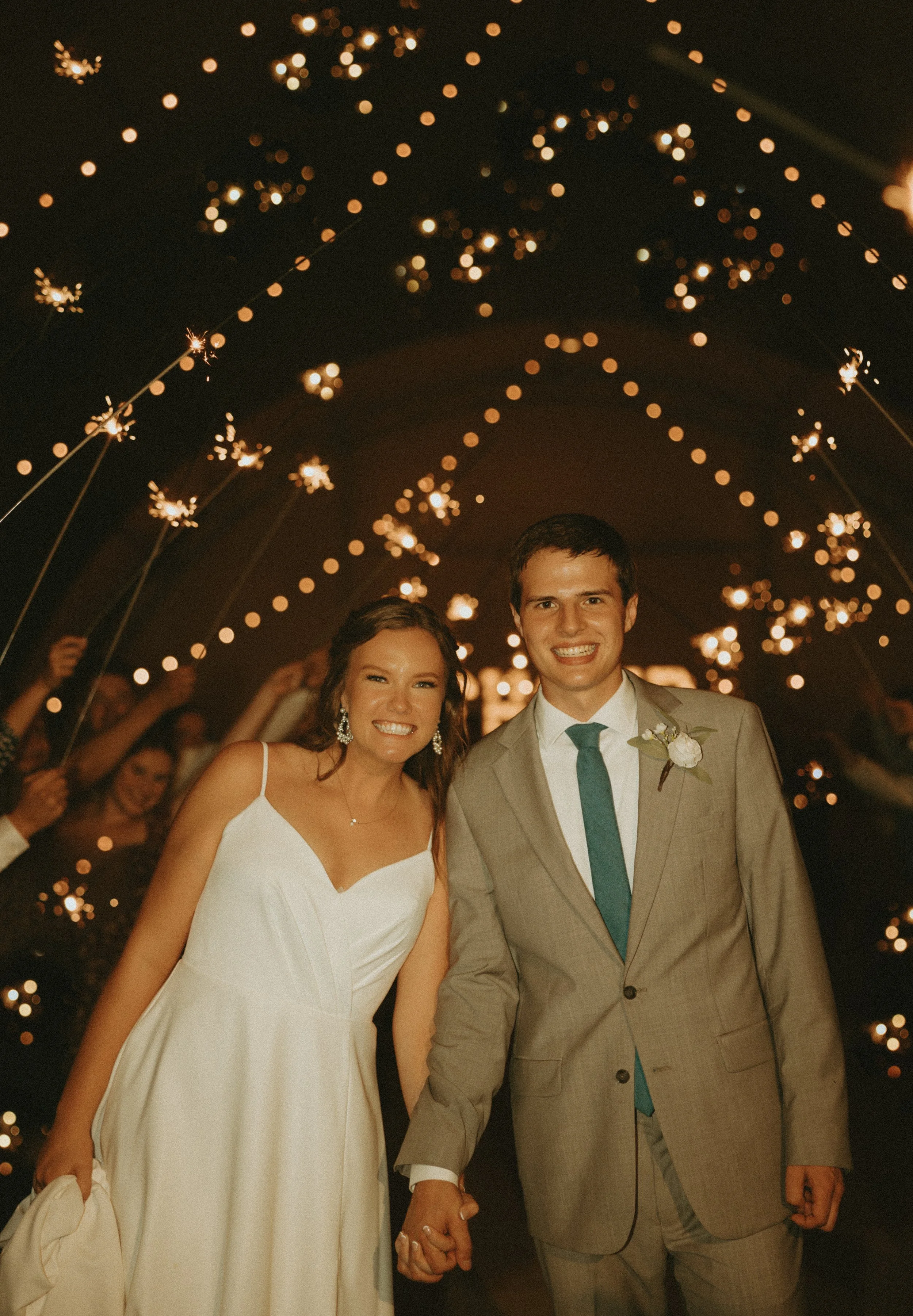 A smiling bride and groom holding hands at night during their wedding celebration, surrounded by guests holding sparklers with string lights overhead.