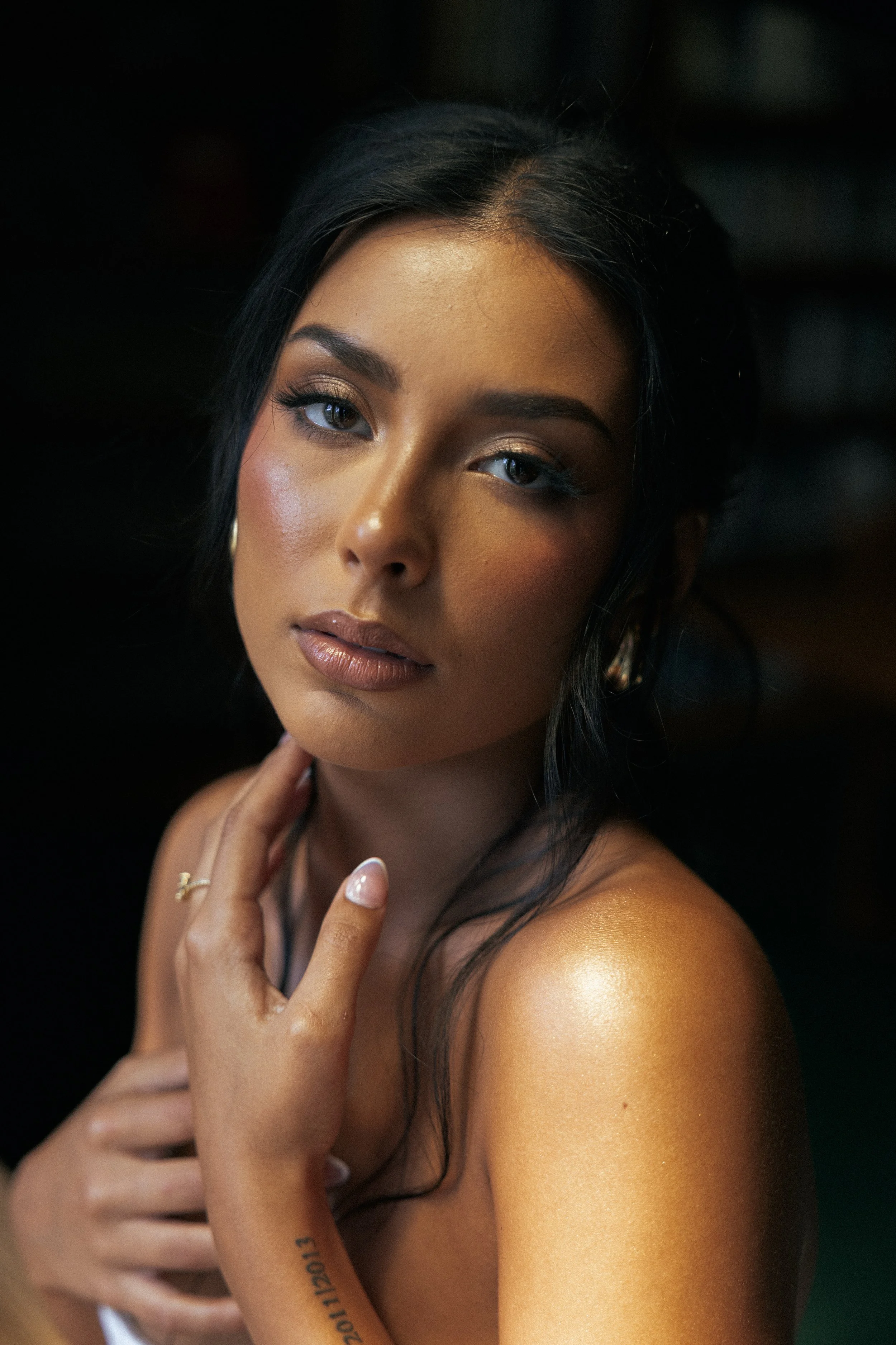 A close-up portrait of a woman with long black hair and tan skin, looking directly at the camera with a neutral expression. She has gold eye shadow, winged eyeliner, and nude lipstick. Her left hand is touching her neck and shoulder, showing a small tattoo on her forearm that reads '2011/12/01' and a ring on her finger.