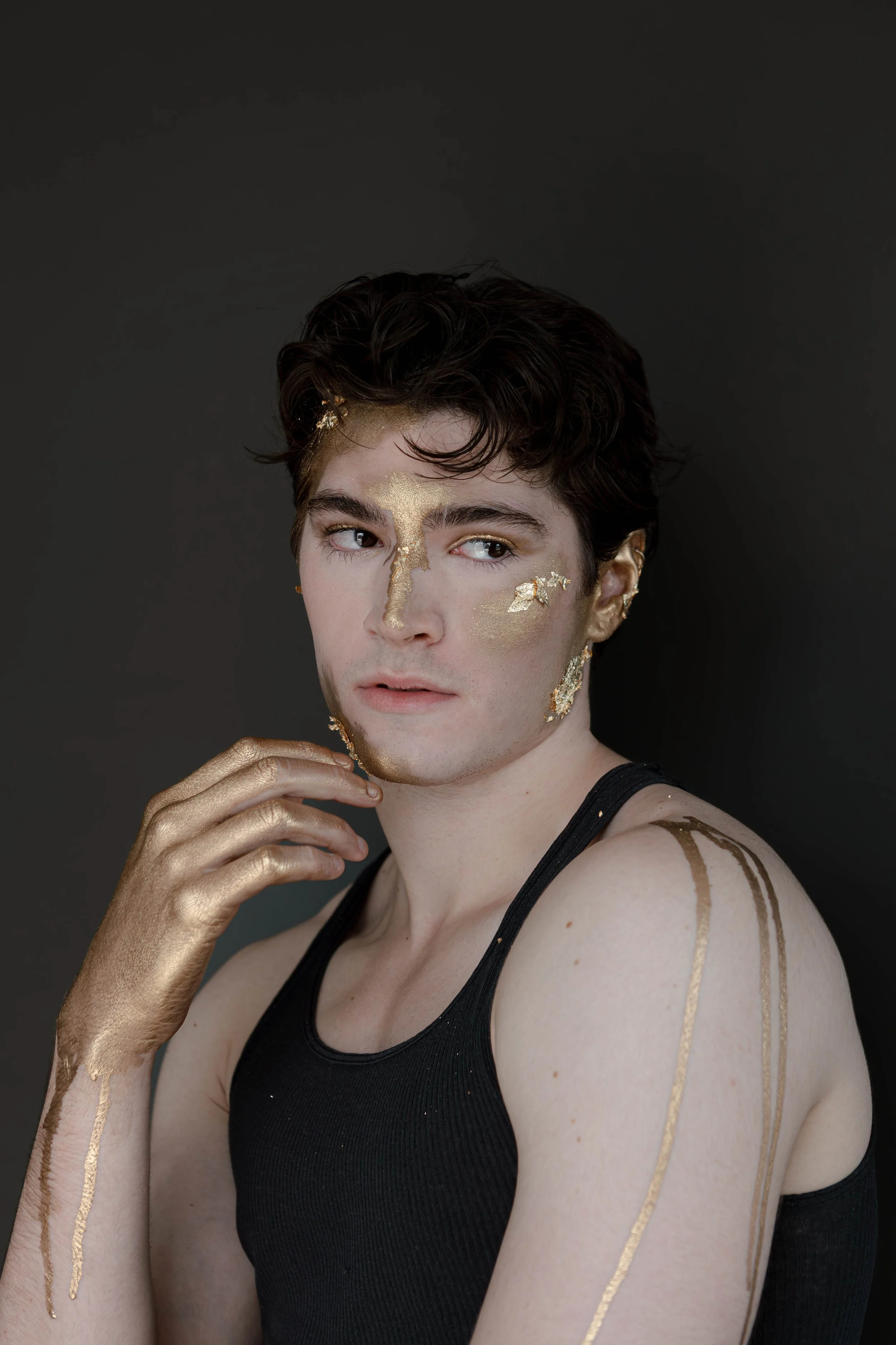 A young person with short dark hair and fair skin poses against a dark background. They are wearing a black tank top. Gold paint is applied to their face, hand, shoulder, and arm, with some areas showing metallic gold streaks and patches on their fac