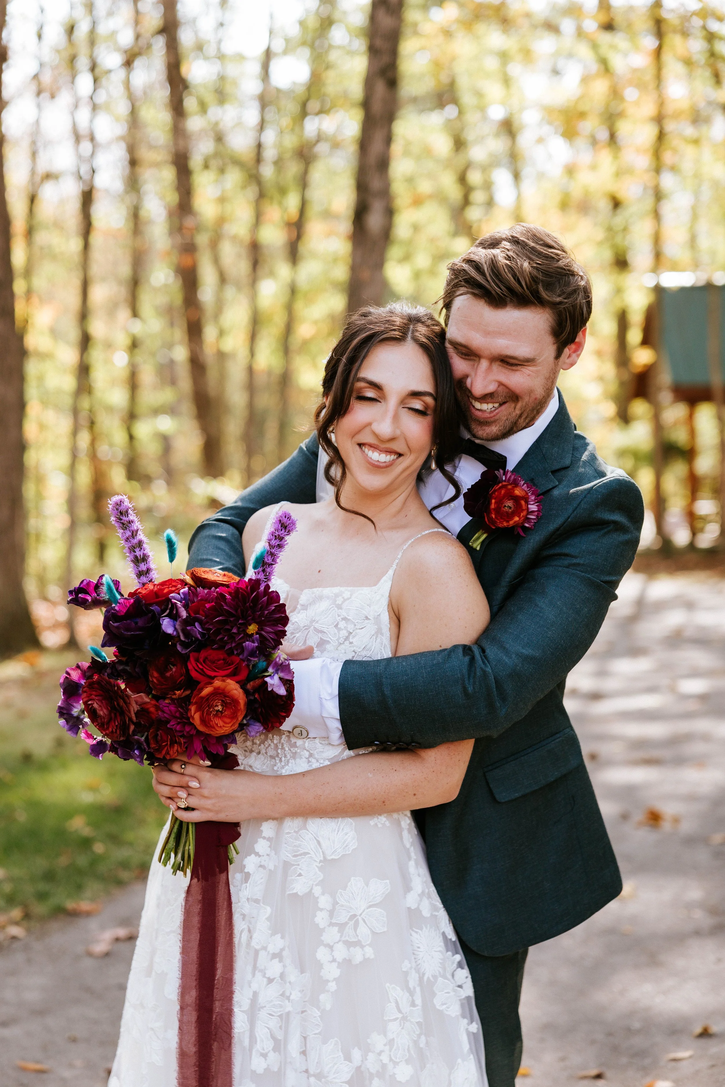 A happy couple on their wedding day, embracing outdoors in a wooded area during autumn. The bride holds a vibrant bouquet of flowers, and they both are smiling joyfully.