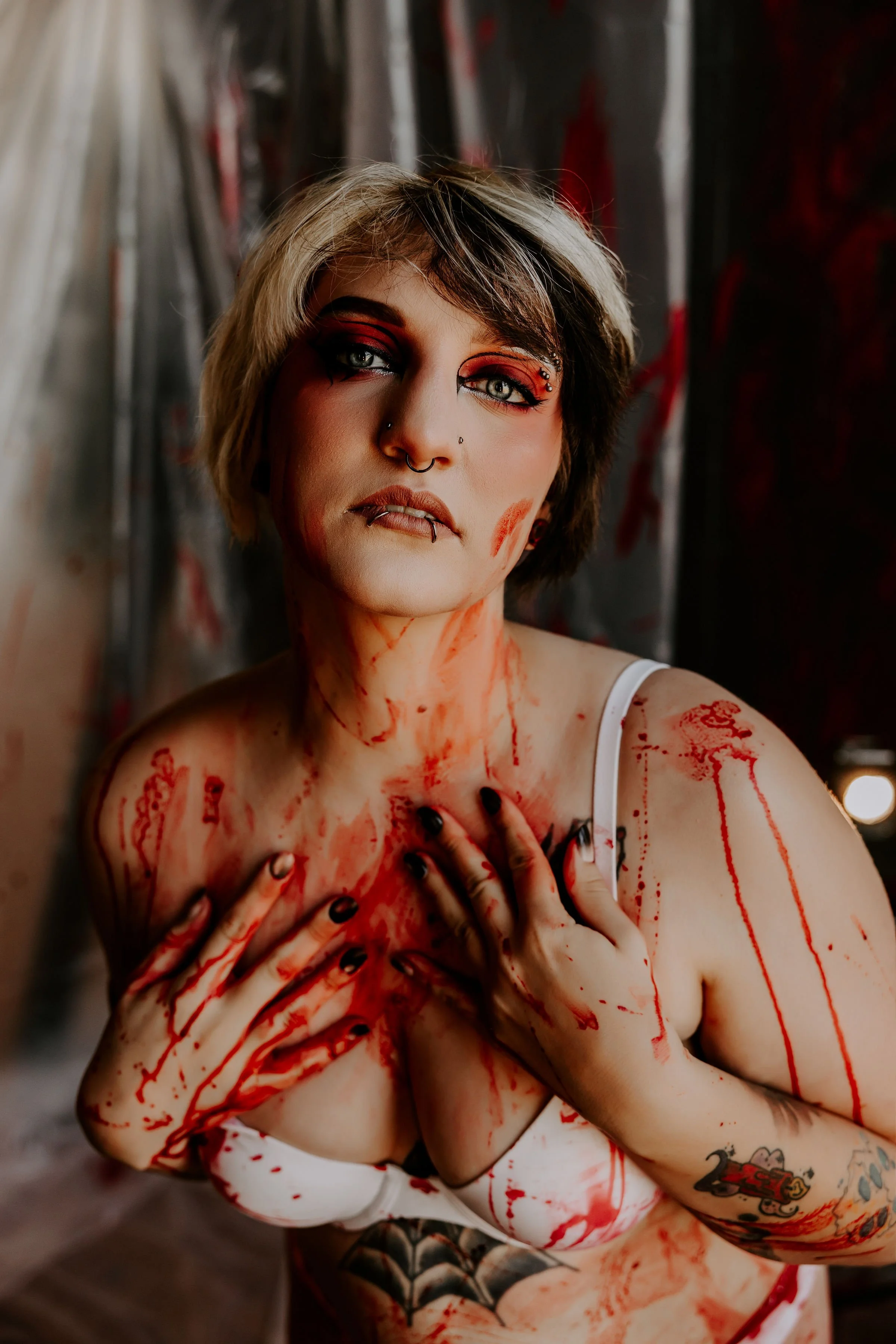 A woman with short hair, makeup, and face piercings. She is covered in fake blood, with a dark background. Her expression is intense as she touches her chest with her hands.