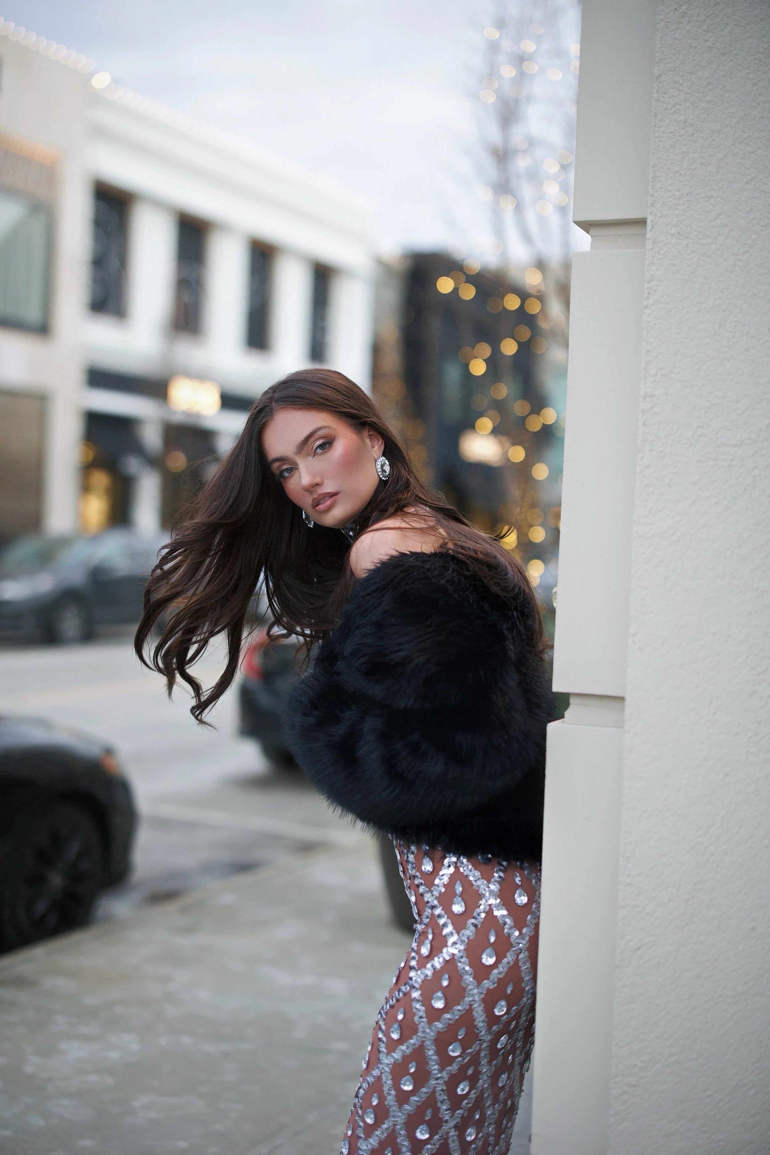 A woman with long dark hair wearing a black fur coat and a sparkly dress with large jewelry is standing outside near a white building, with blurred city scene and festive lights in the background during evening or dusk.