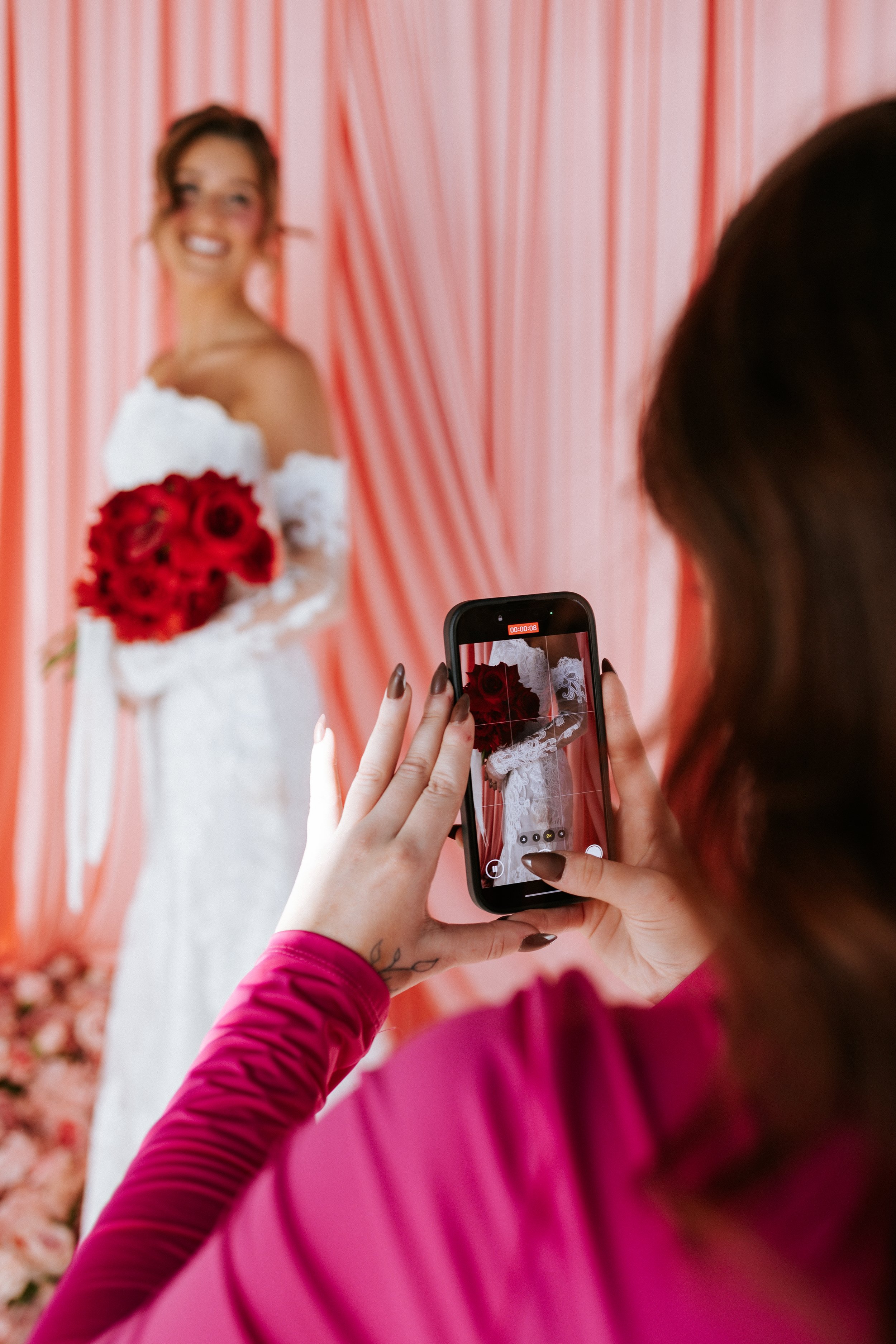 A woman in a magenta long-sleeve top taking a photo of another woman in a white dress holding a bouquet of red roses against a peach and pink striped curtain background.