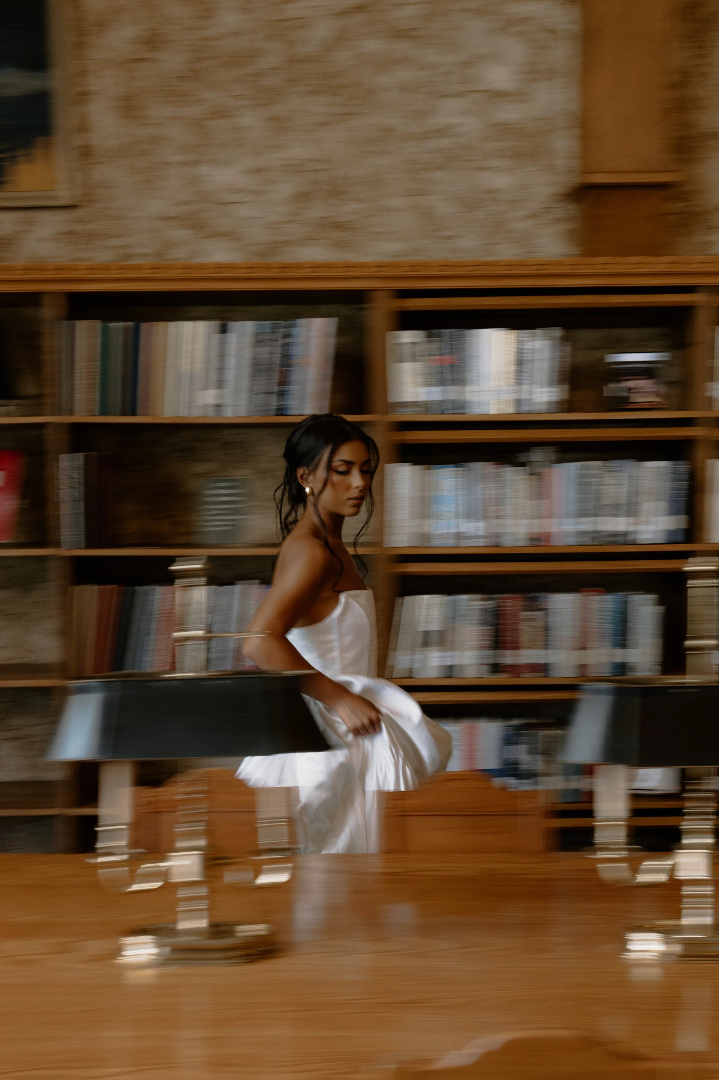 A woman in a white dress sitting in a room with wooden shelves filled with books, appearing blurry due to motion.