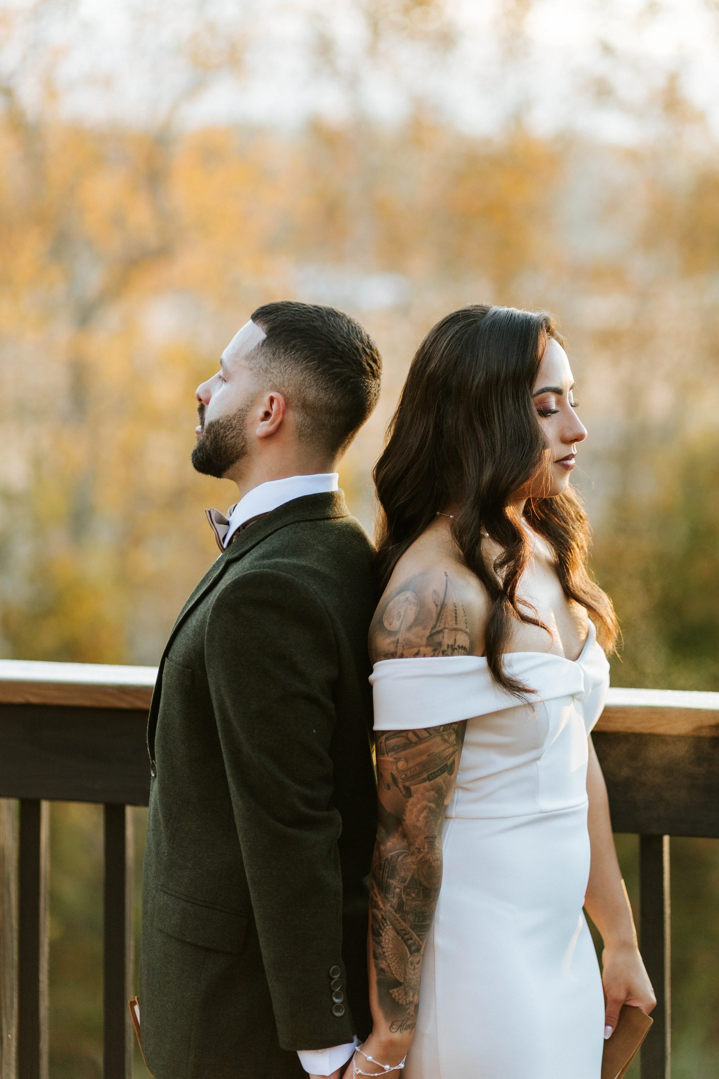 A man with a beard in a green suit and a woman with long dark hair and tattoos on her arm are standing back to back on a wooden deck outside, with a blurred autumnal background.