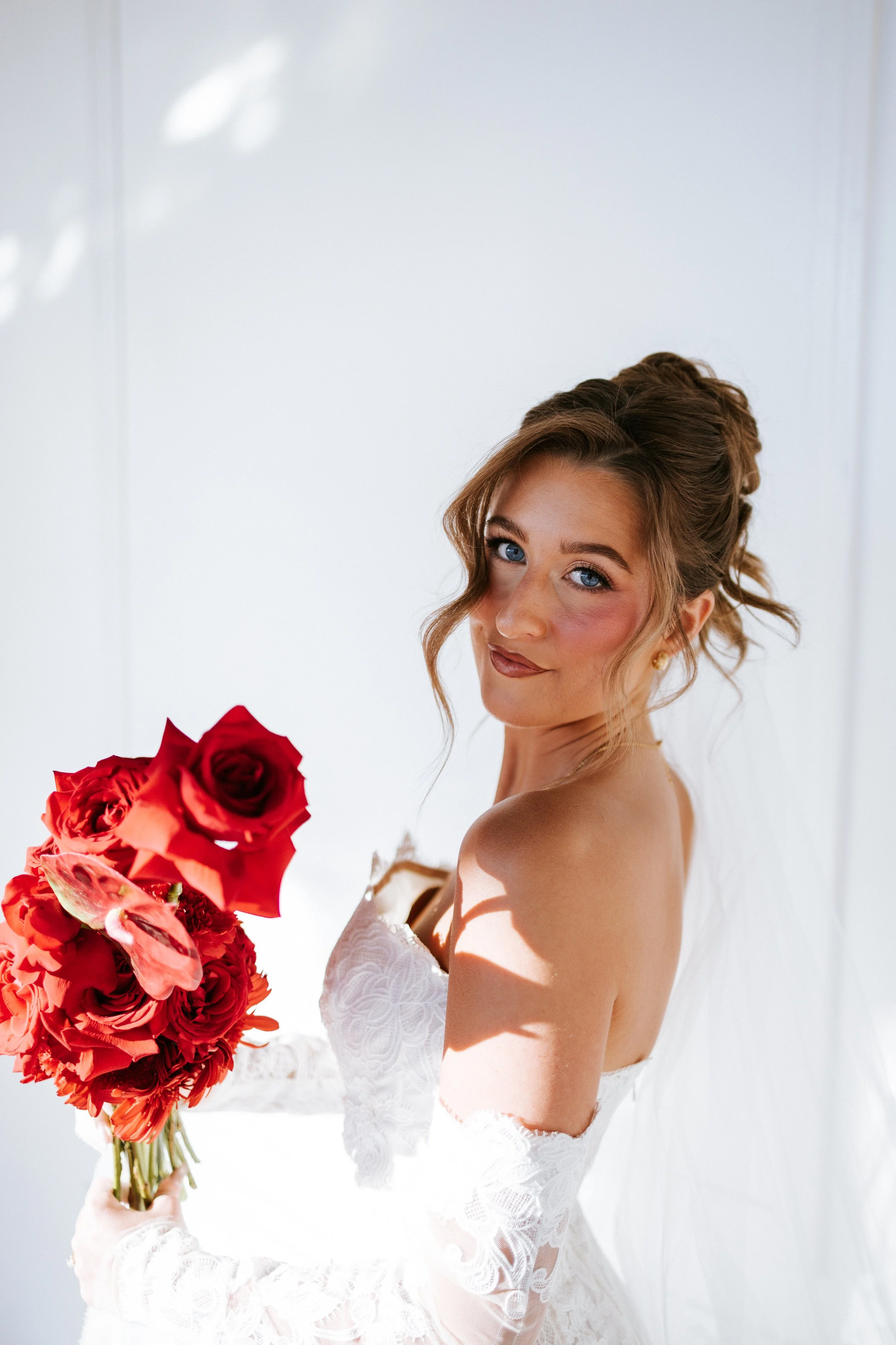 A young woman in a white lace dress holding a bouquet of red and orange roses, standing against a plain white background.