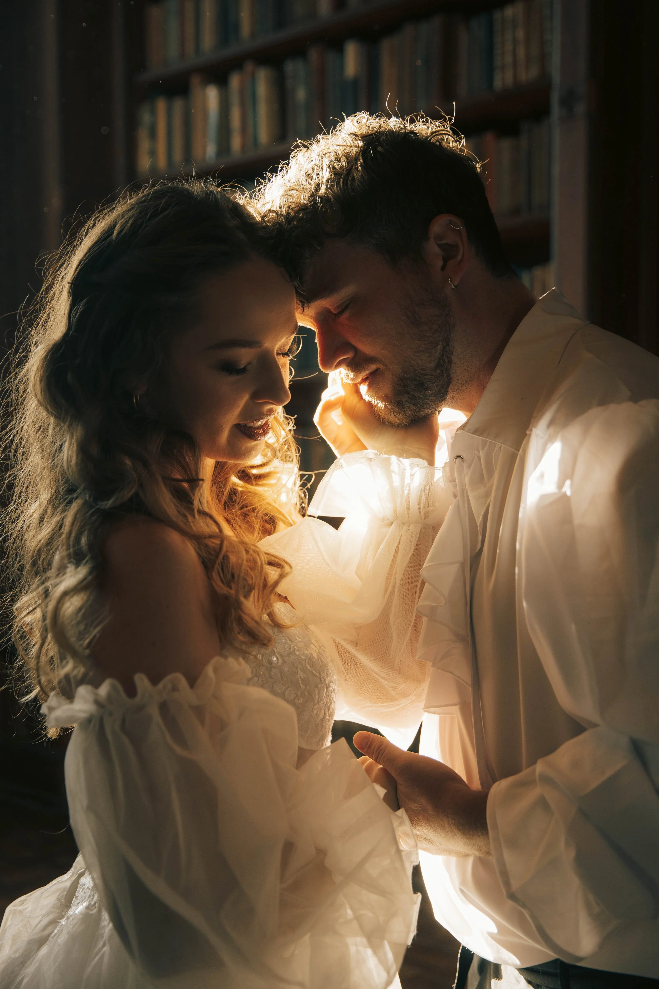A couple with their foreheads touching, smiling and holding hands in a warmly lit room with bookshelves in the background.
