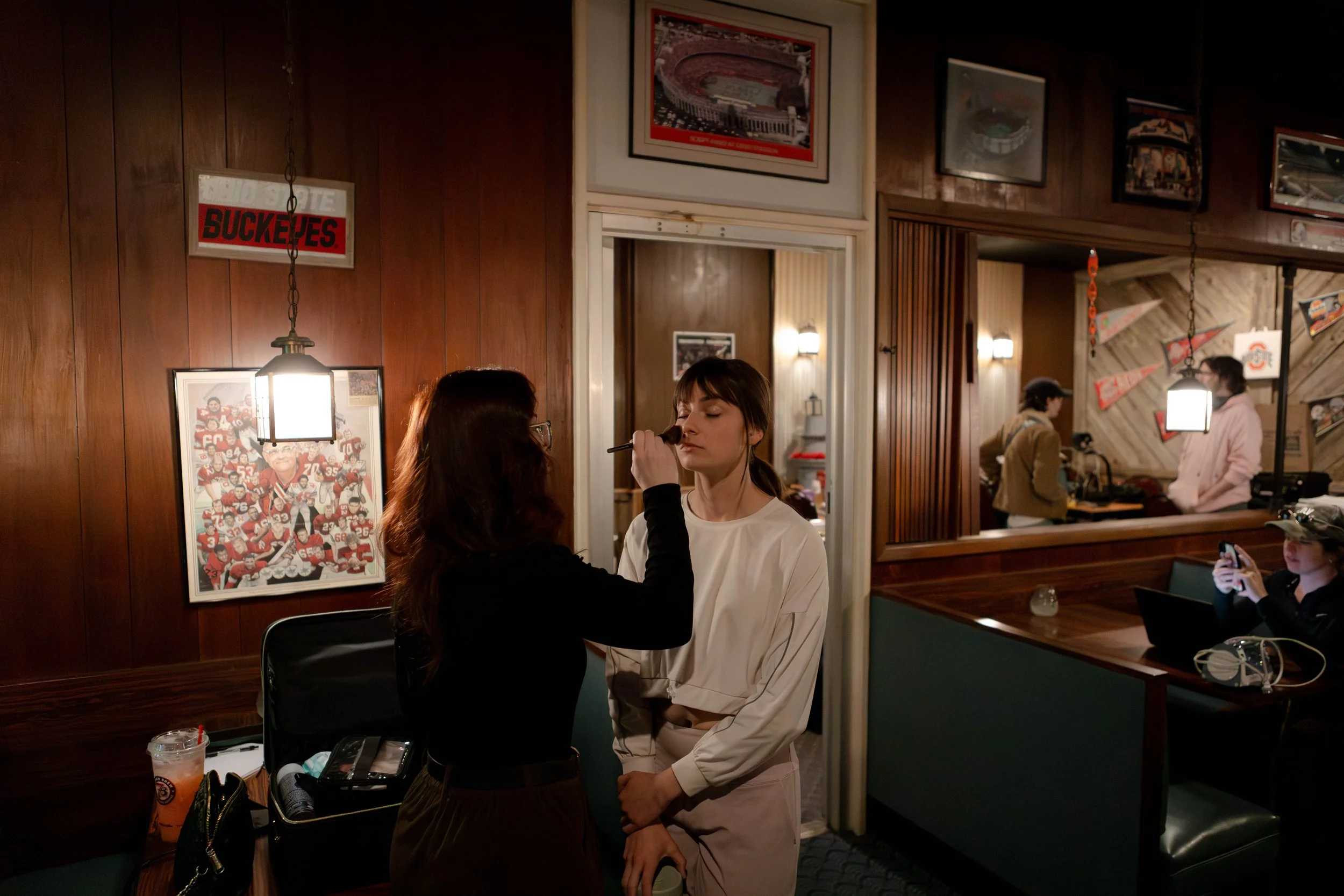 Two women are in a restaurant. One is applying makeup to the other, who is standing with eyes closed. The restaurant has wood-paneled walls, sports-themed posters, and several other patrons, including a woman taking a selfie.
