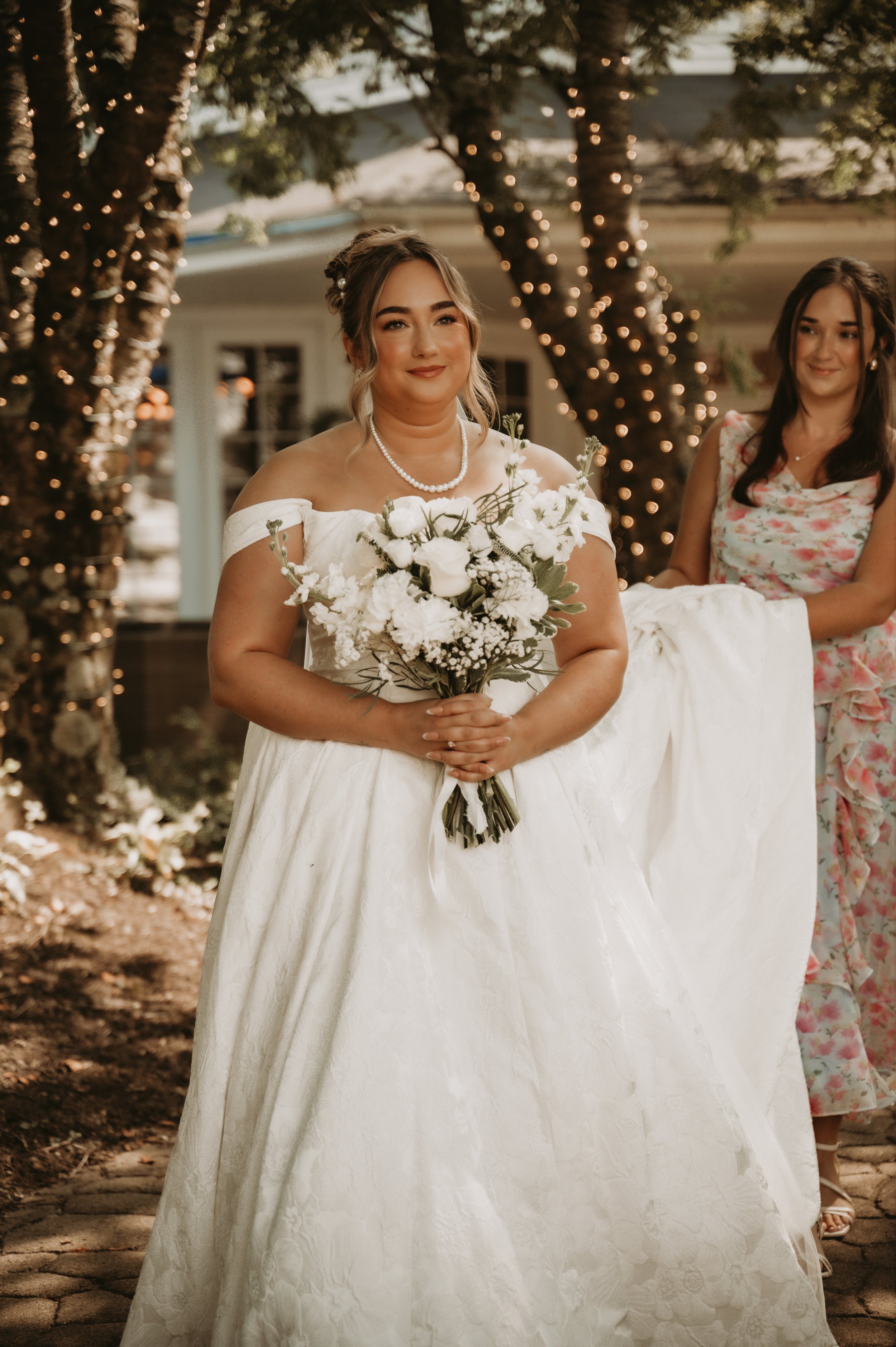 A bride in a white off-the-shoulder wedding dress holding a bouquet of white flowers, standing outdoors under string lights wrapped around trees, with another woman in a floral dress nearby.