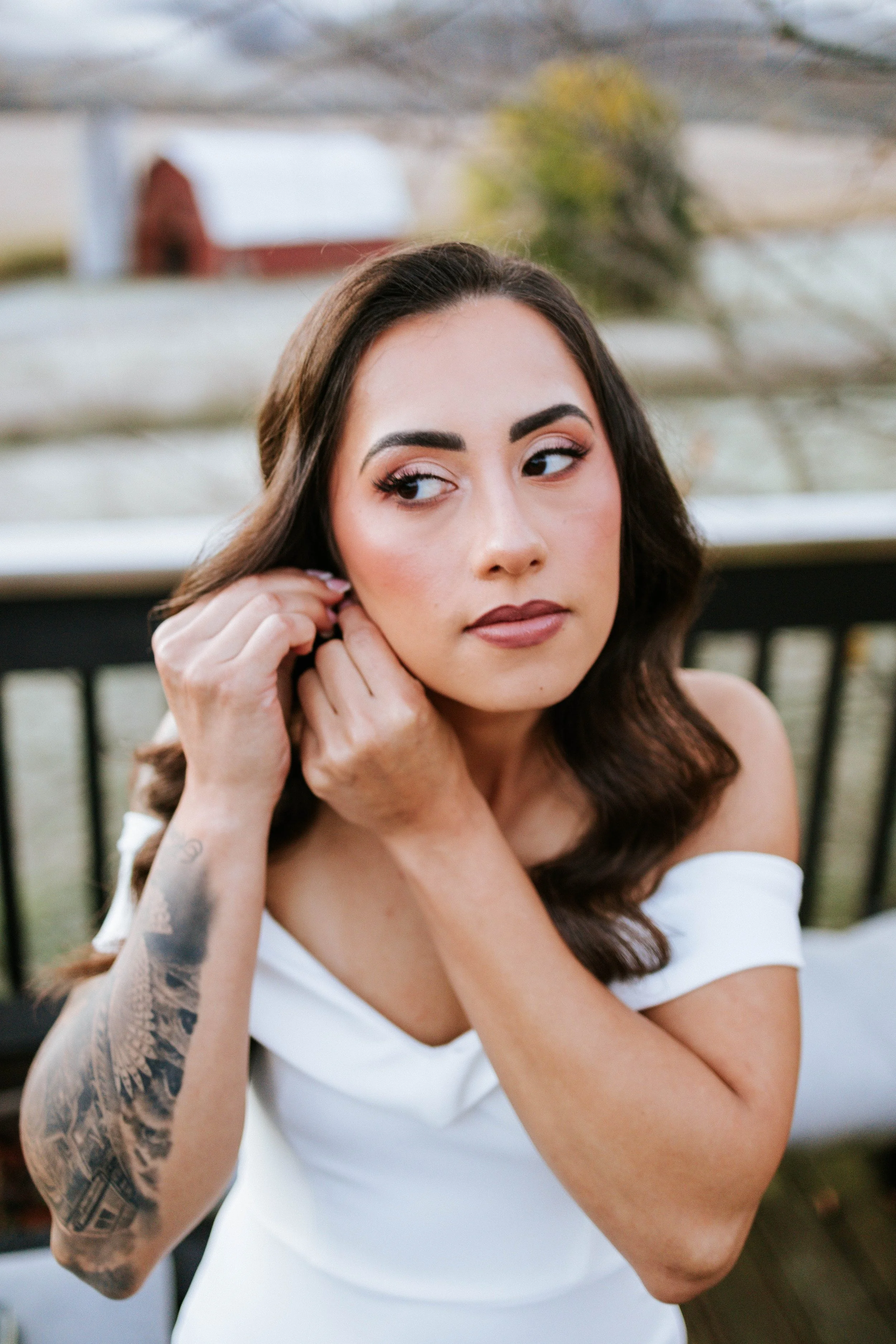 A woman with dark brown hair and tattoos on her arm is adjusting her earring while sitting outdoors. She is wearing a white off-the-shoulder top and has makeup with pink eyeshadow and lipstick. The background features a blurred view of a house and tr