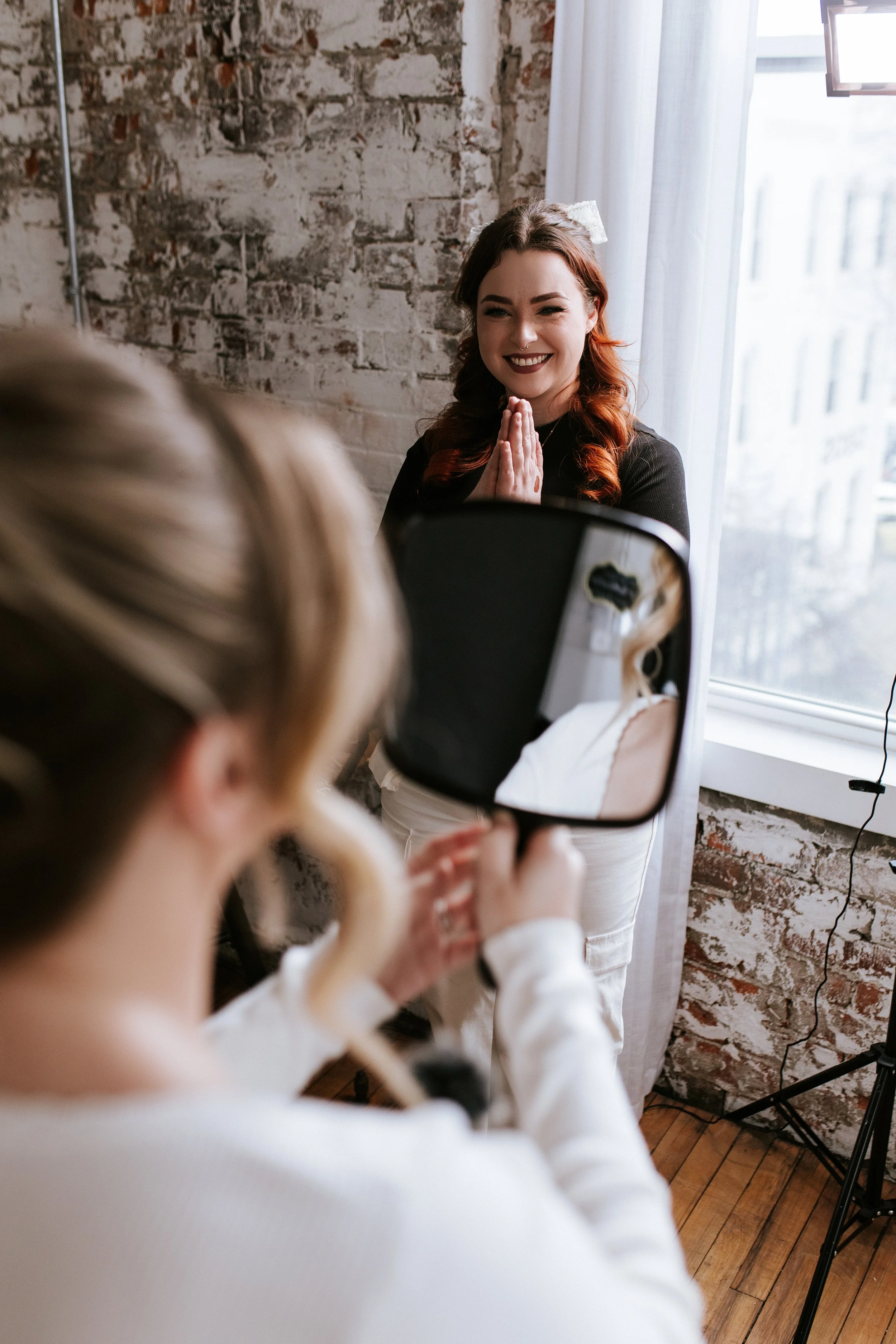A woman with red hair and a big white bow in her hair is smiling with her hands clasped together near her face. She is looking at her reflection in a mirror held by another person. The setting appears to be a room with a brick wall, wooden floor, and a window with white curtains.