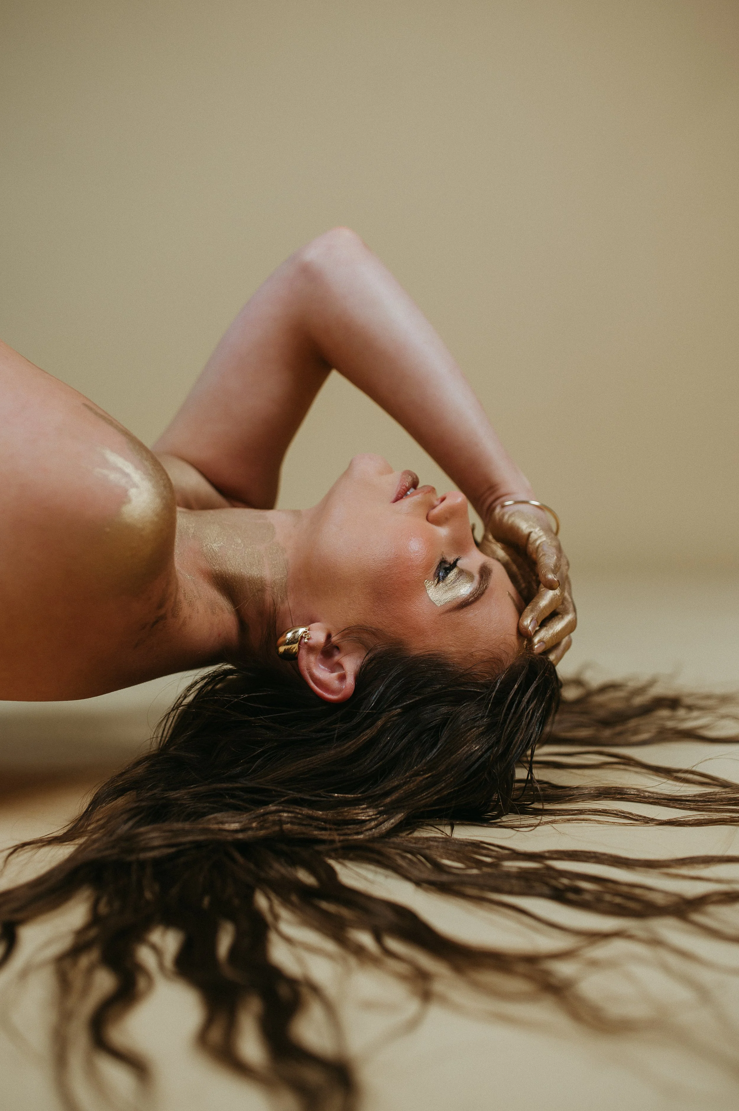 A woman lying on her back with long, wavy brown hair spread out on the floor, holding her head with one hand and looking upward, with gold and neutral makeup and gold jewelry.