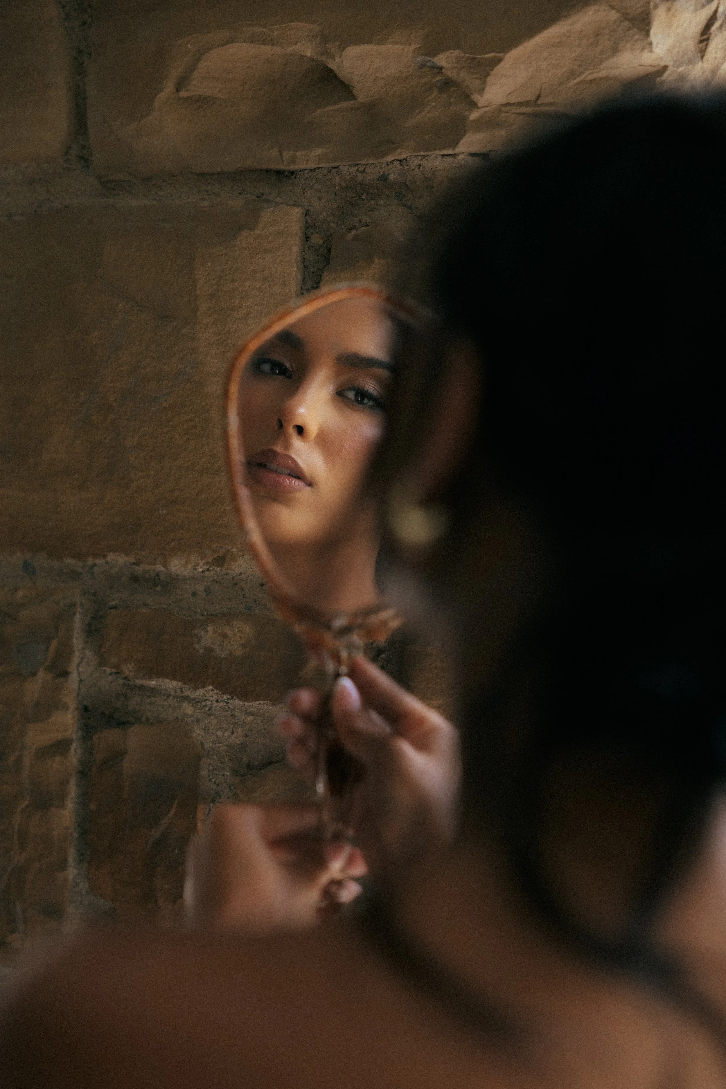 A woman looking at her reflection in a small handheld mirror. She has light makeup and is against a brick wall.
