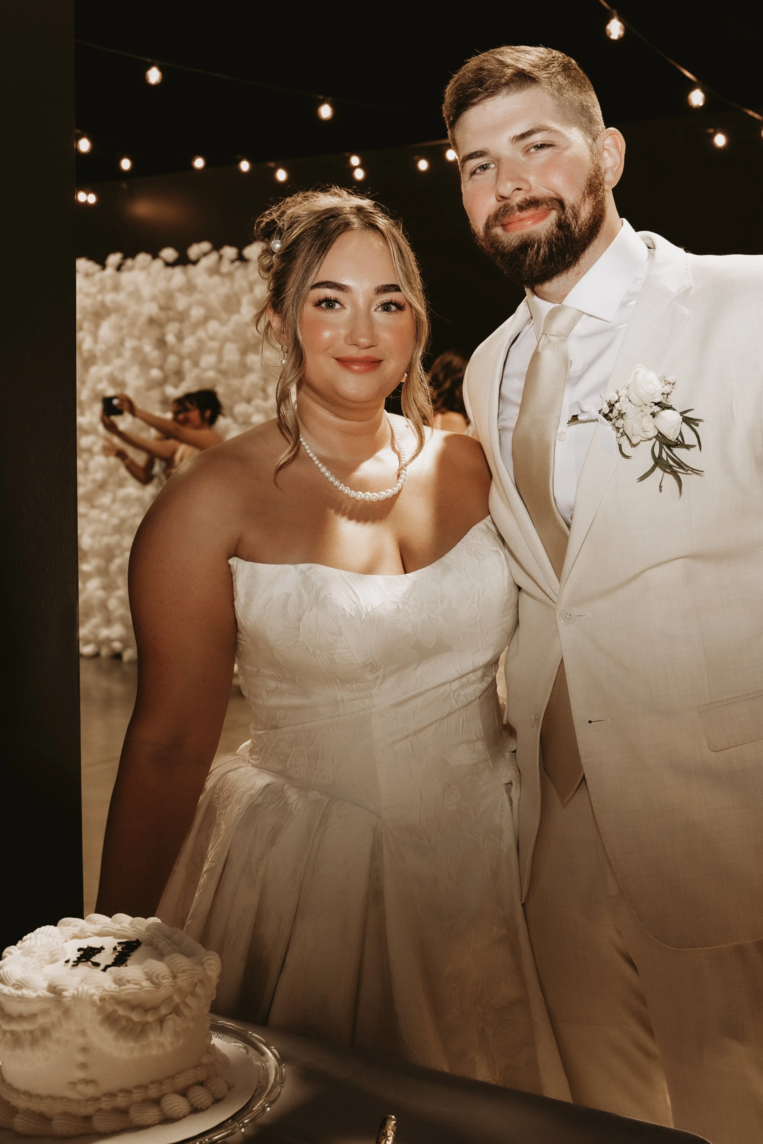 A bride and groom at their wedding reception, standing near a decorated table with a cake. The bride wears a strapless wedding dress and pearl necklace, and the groom is in a white suit with a boutonnière. Background features string lights and a whit