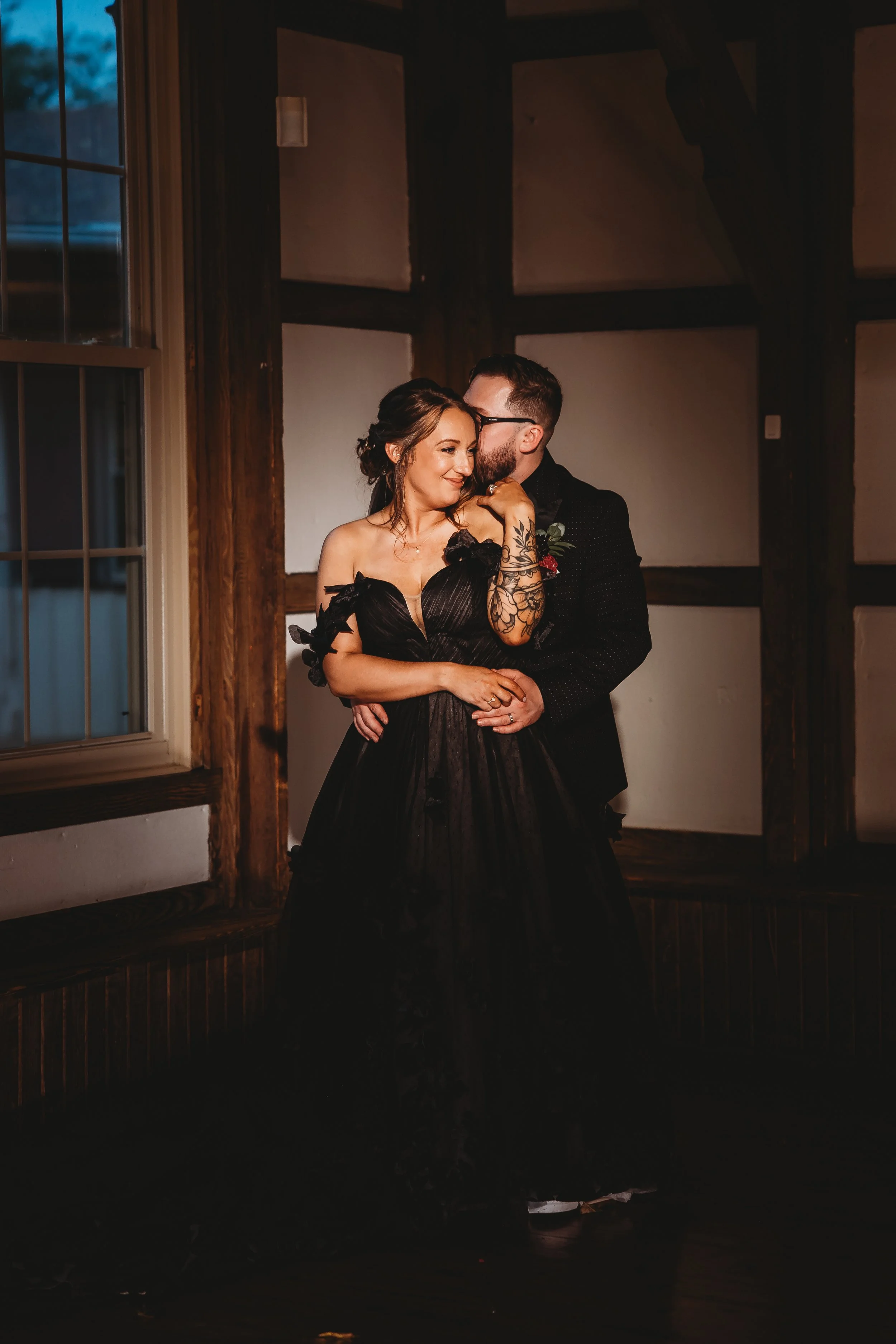 A couple, dressed in formal black attire, is dancing closely in a dimly lit room with wooden walls and large windows.