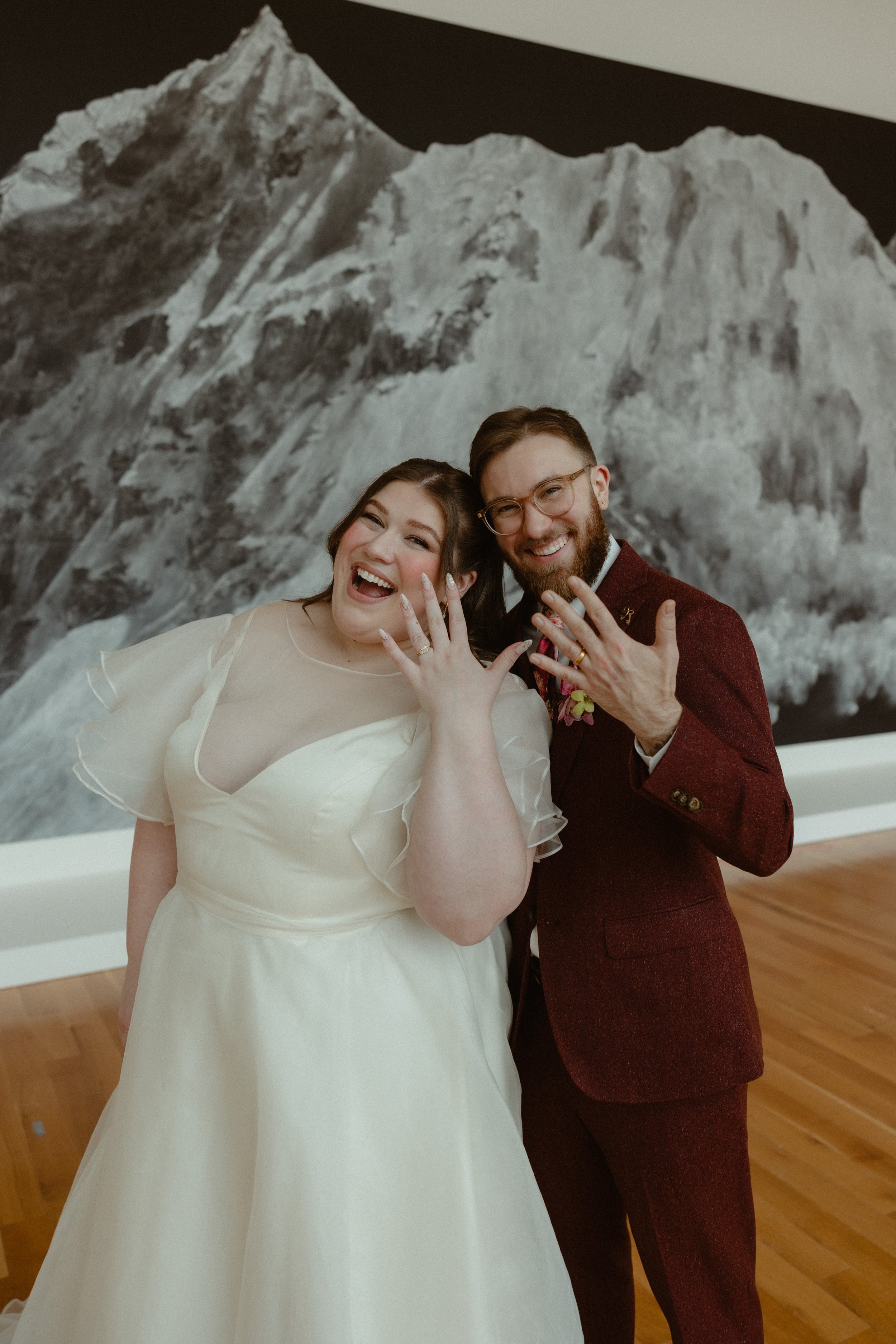 A happy bride and groom showing off their wedding rings, standing in front of a large mountain photograph at their wedding celebration.