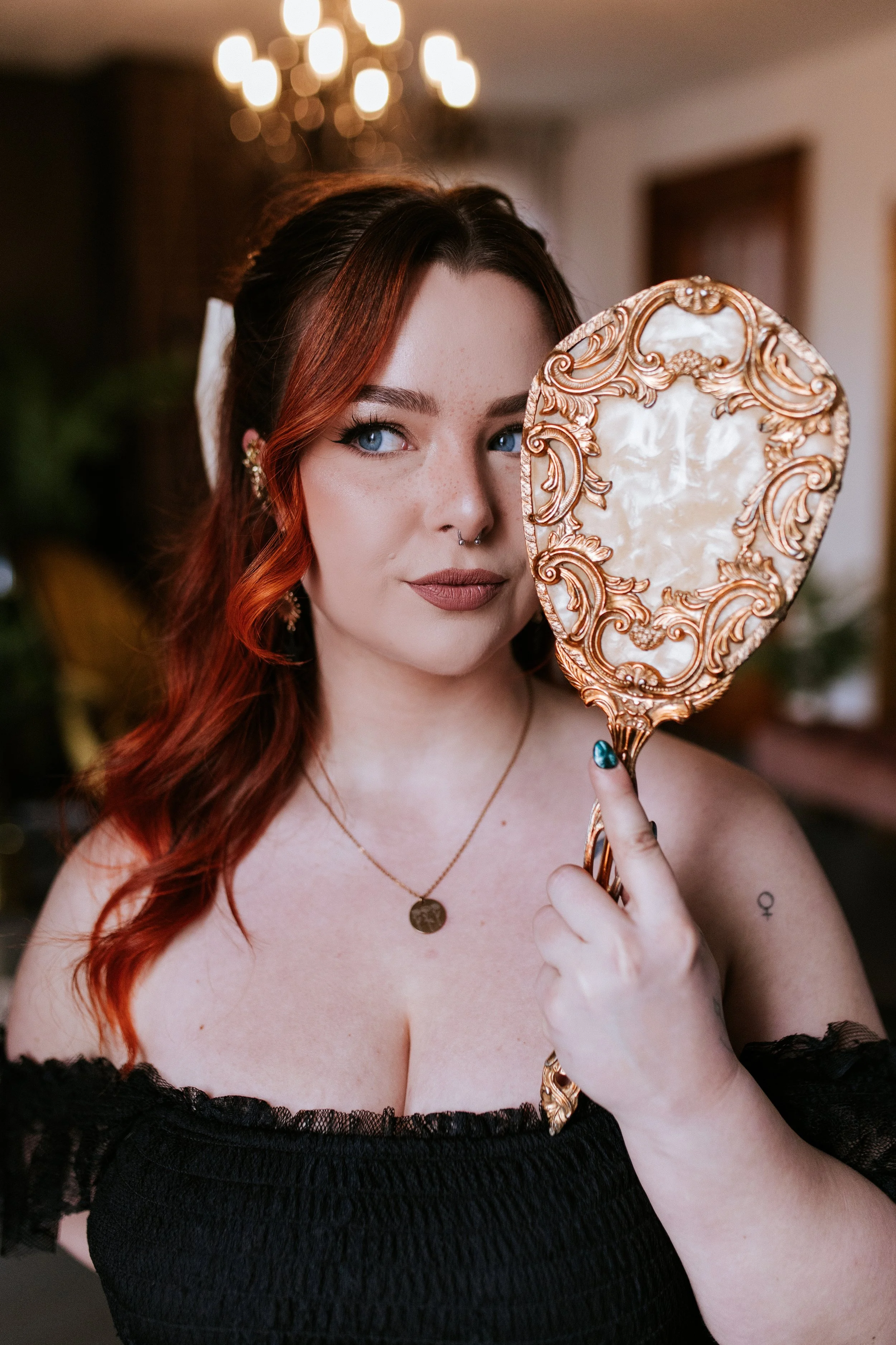 A woman with red hair and blue eyes holds a ornate hand mirror with a gold frame and mother-of-pearl back, partially covering one eye, wearing black lace off-shoulder top and gold jewelry.