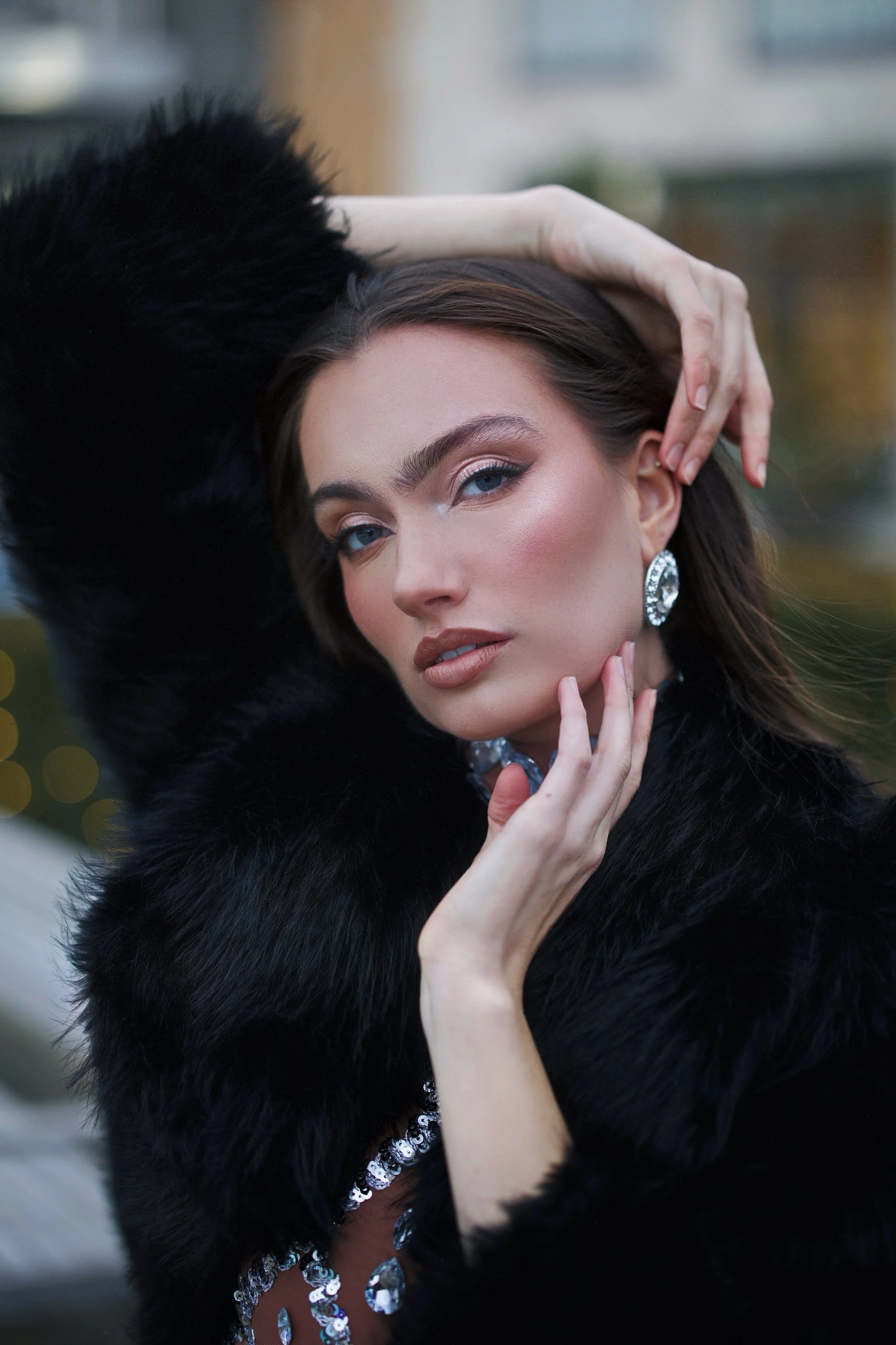 Close-up of a woman with blue eyes, wearing sparkling earrings, a dark fur coat, and a dress adorned with sequins and large gemstones, posed with her hand on her head.