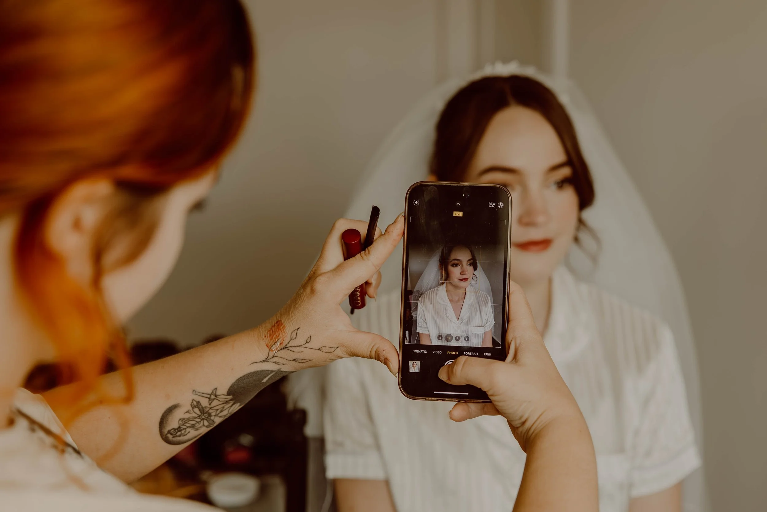 A woman with tattoos on her arm takes a photo with her phone of another woman in a white dress and veil, who is looking at her reflection in a mirror.