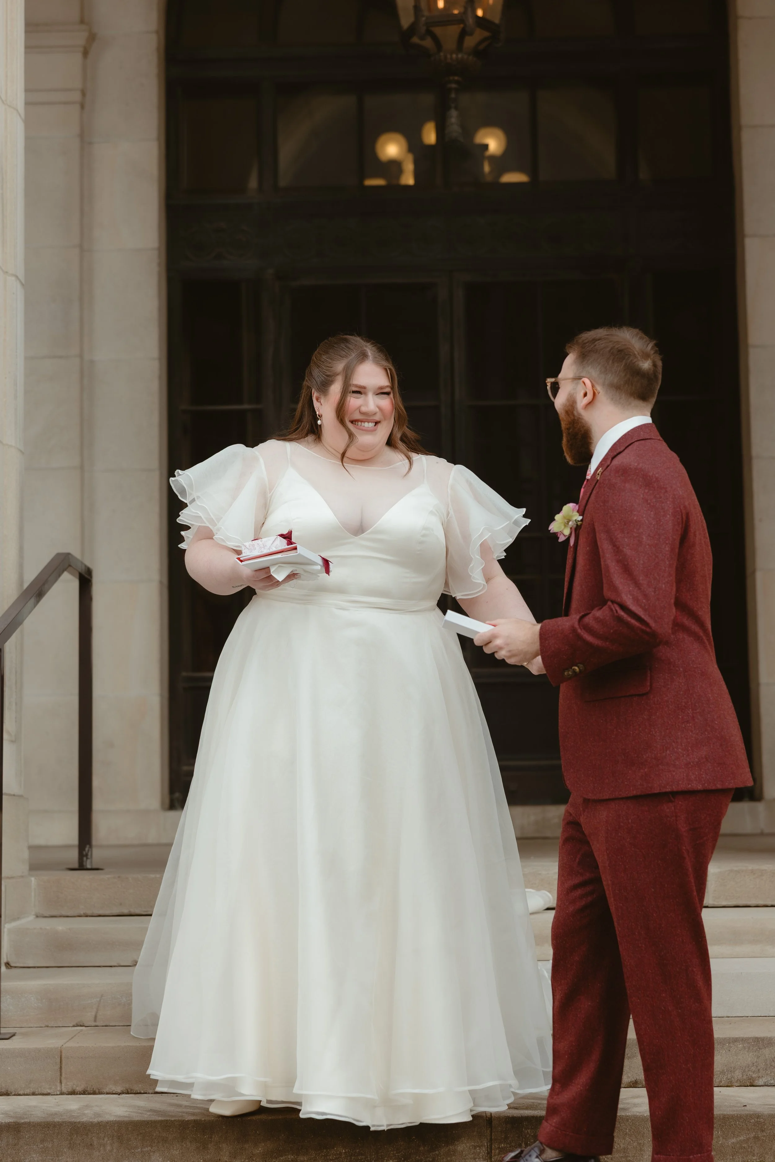 A woman in a white wedding dress and a man in a red suit are smiling and holding hands on the steps of a building during a wedding ceremony.