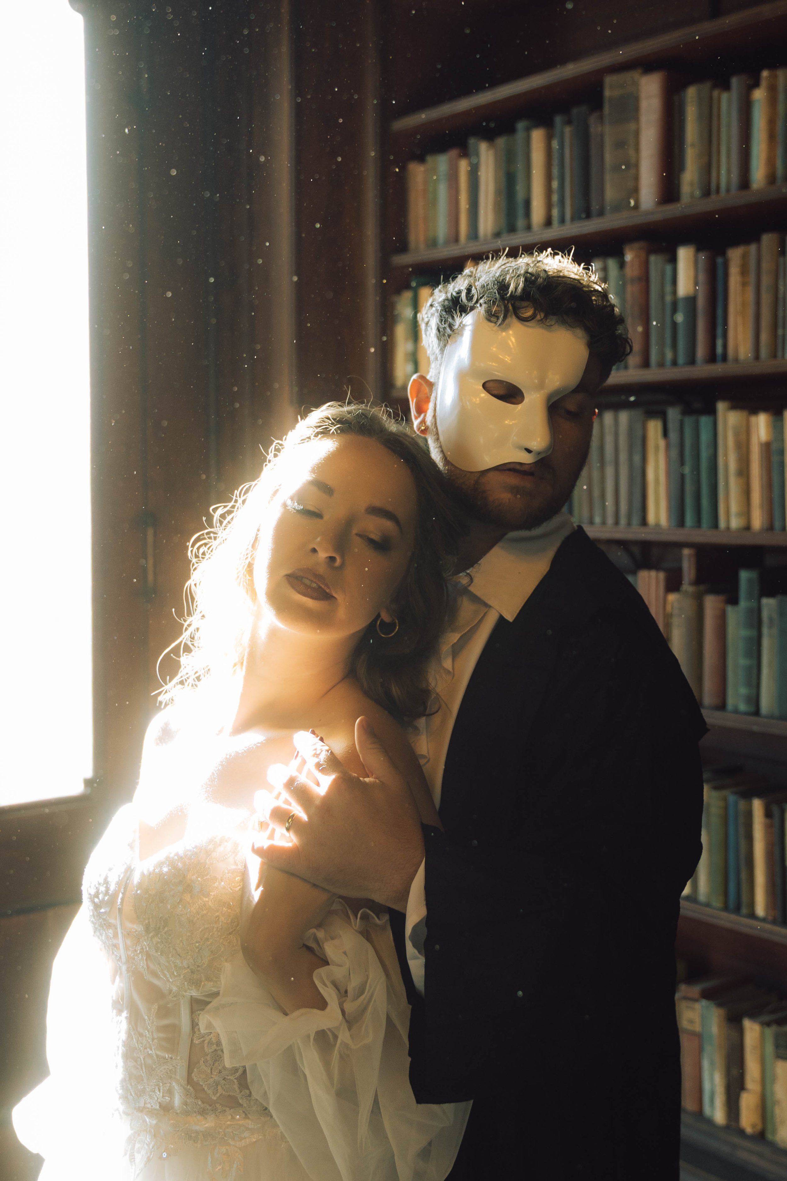 A man wearing a white mask is standing with a woman with light skin and long, wavy hair. The woman with closed eyes is dressed in a white lacy dress and is embracing the man. They are indoors, near a large window with bright sunlight streaming in, in