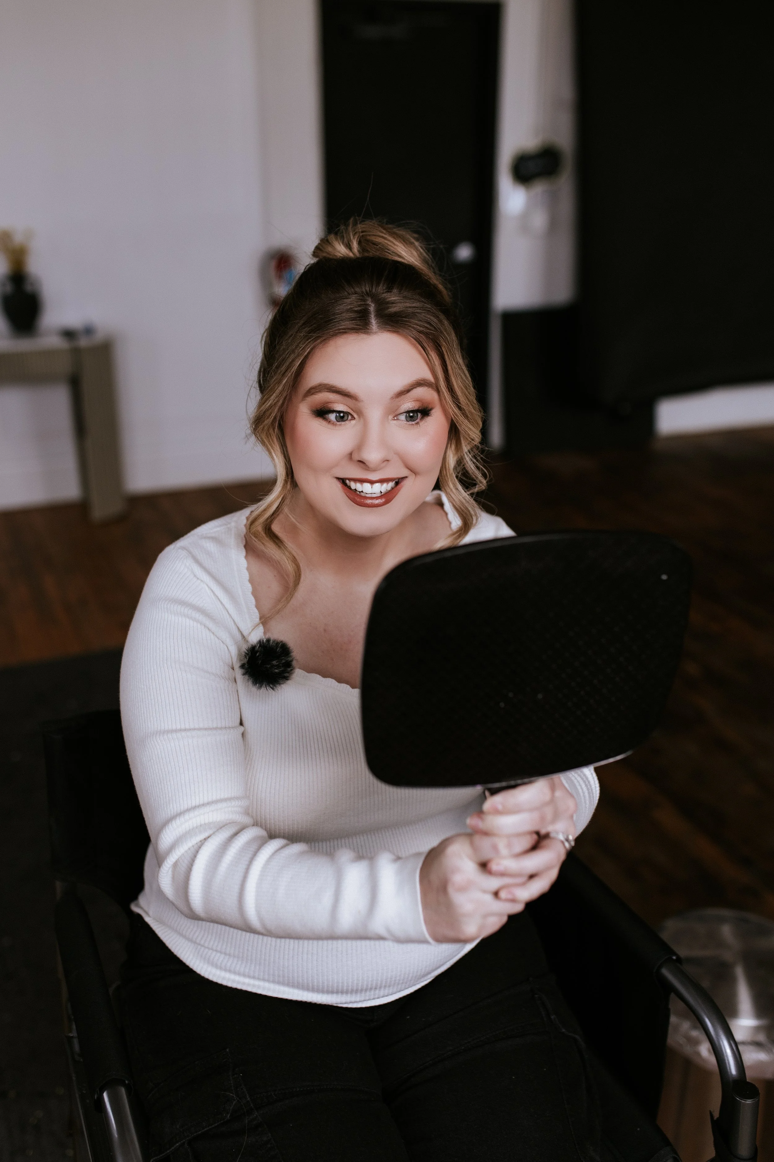 A woman sitting in a wheelchair, holding a mirror, smiling with makeup on, in a cozy room.