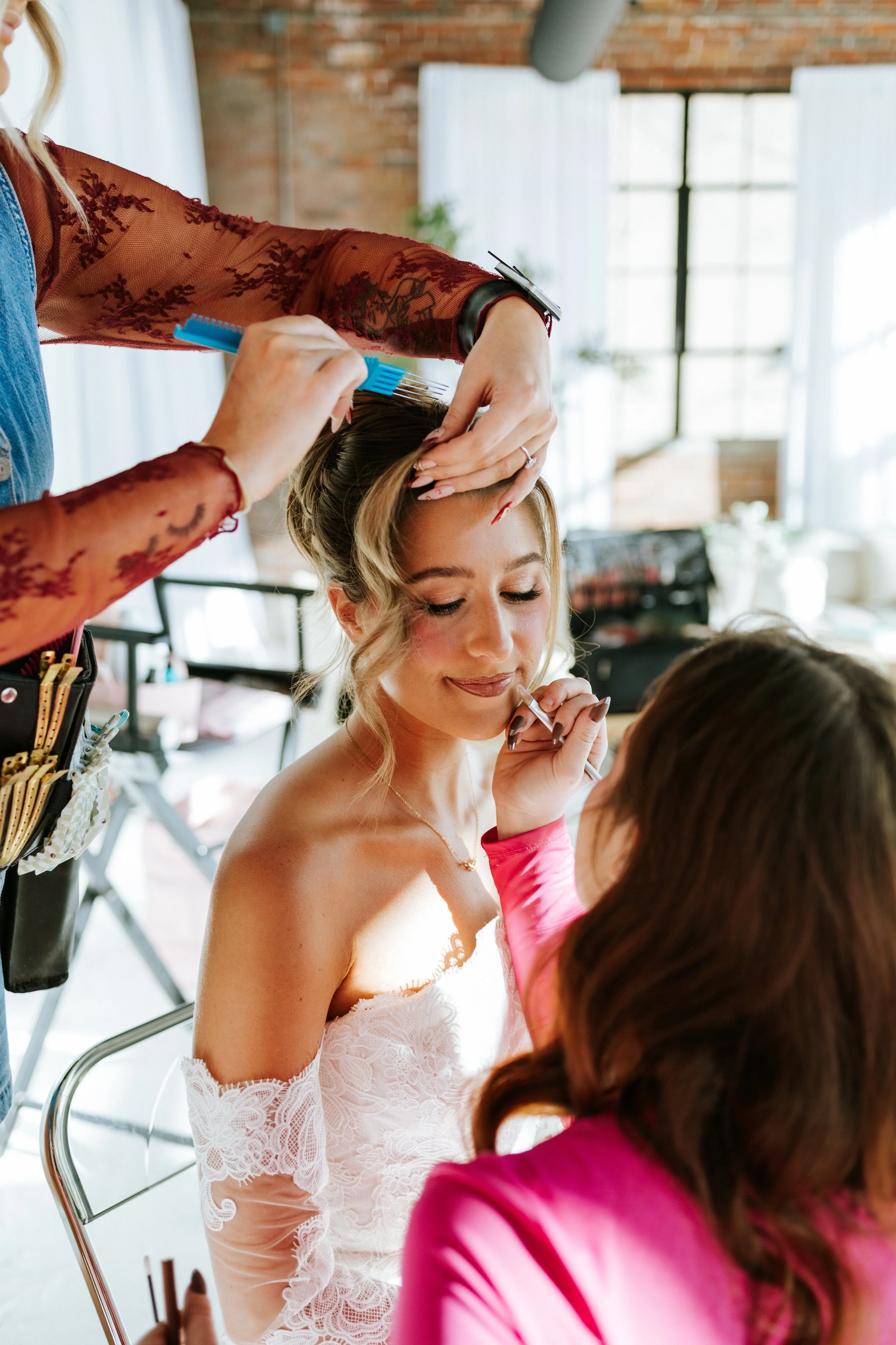 A woman in a white lace dress is getting her makeup done while another woman in a pink jacket is doing her makeup in a bright, airy room with large windows and brick walls.