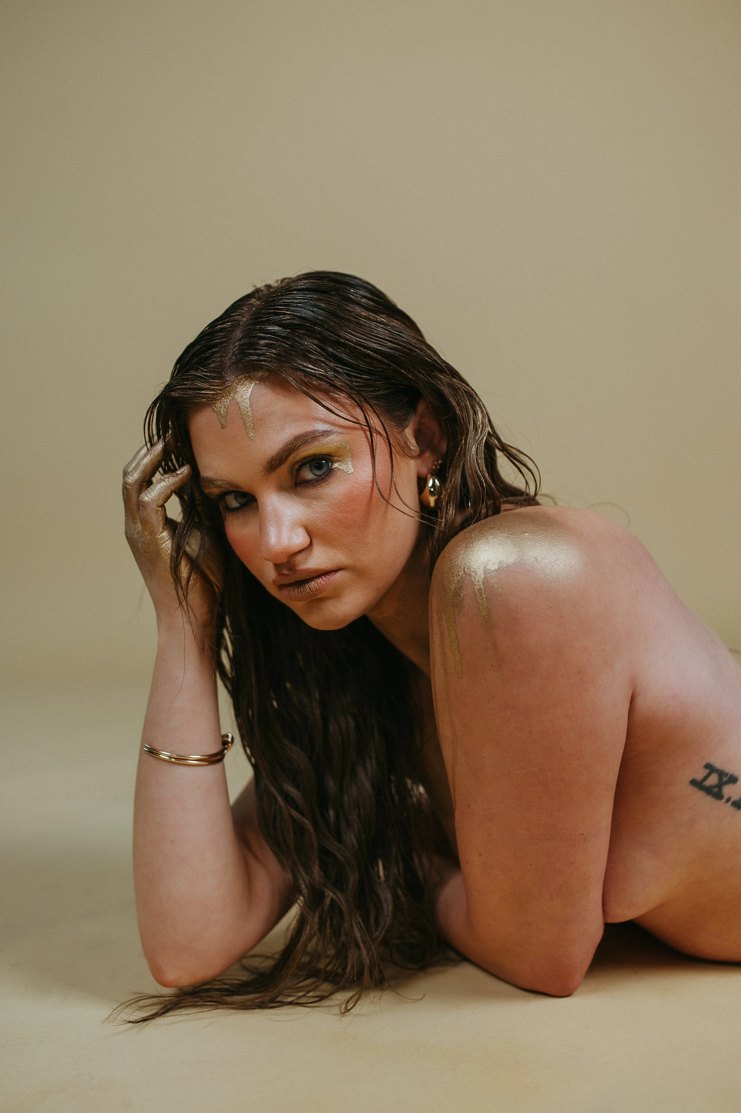 A woman with wet dark hair and gold paint on her face and shoulder, lying on the floor with a neutral expression, wearing jewelry and a tattoo on her arm, against a plain beige background.