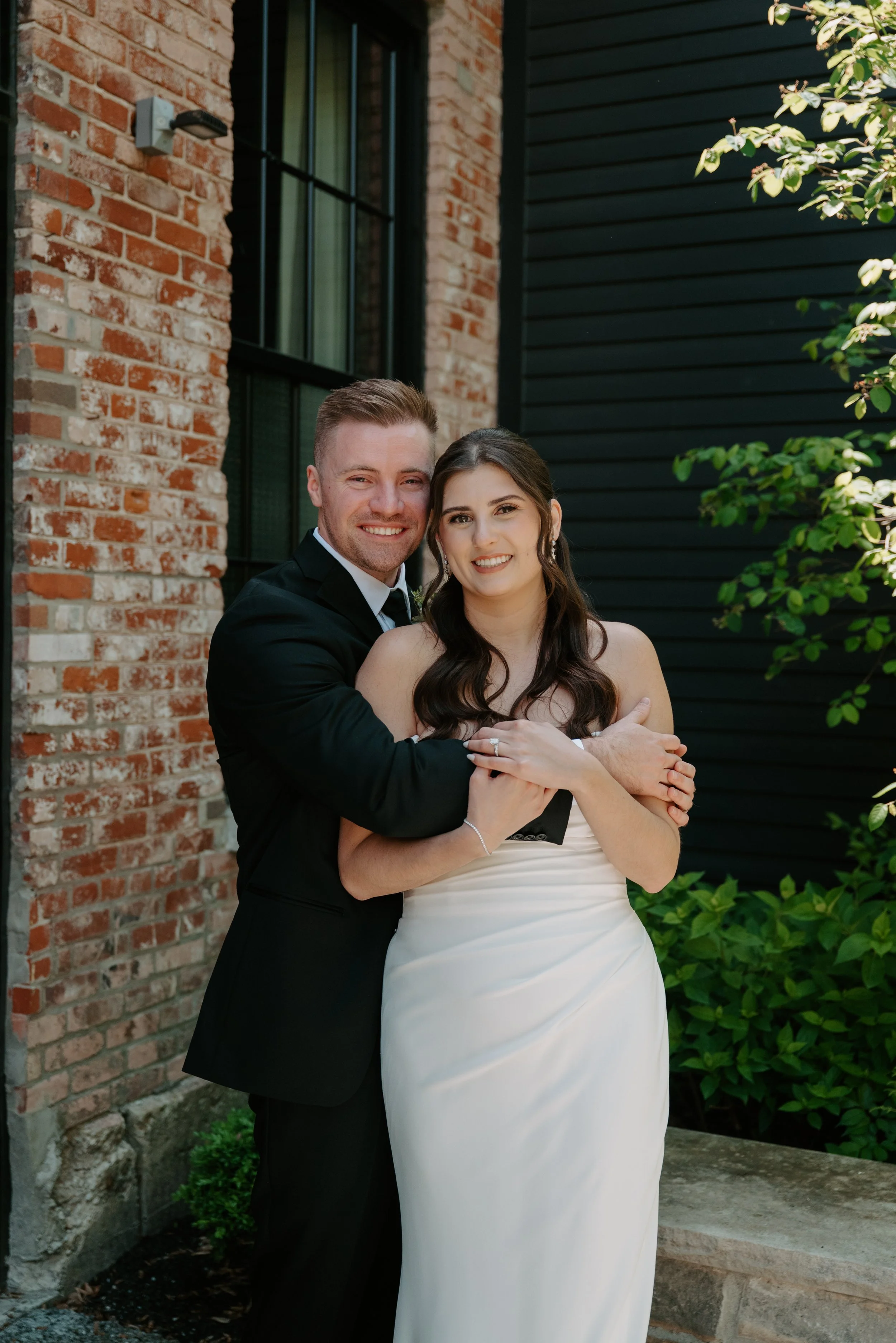 A smiling man and woman embracing outdoors next to a brick and black siding wall, with green foliage on the right side.