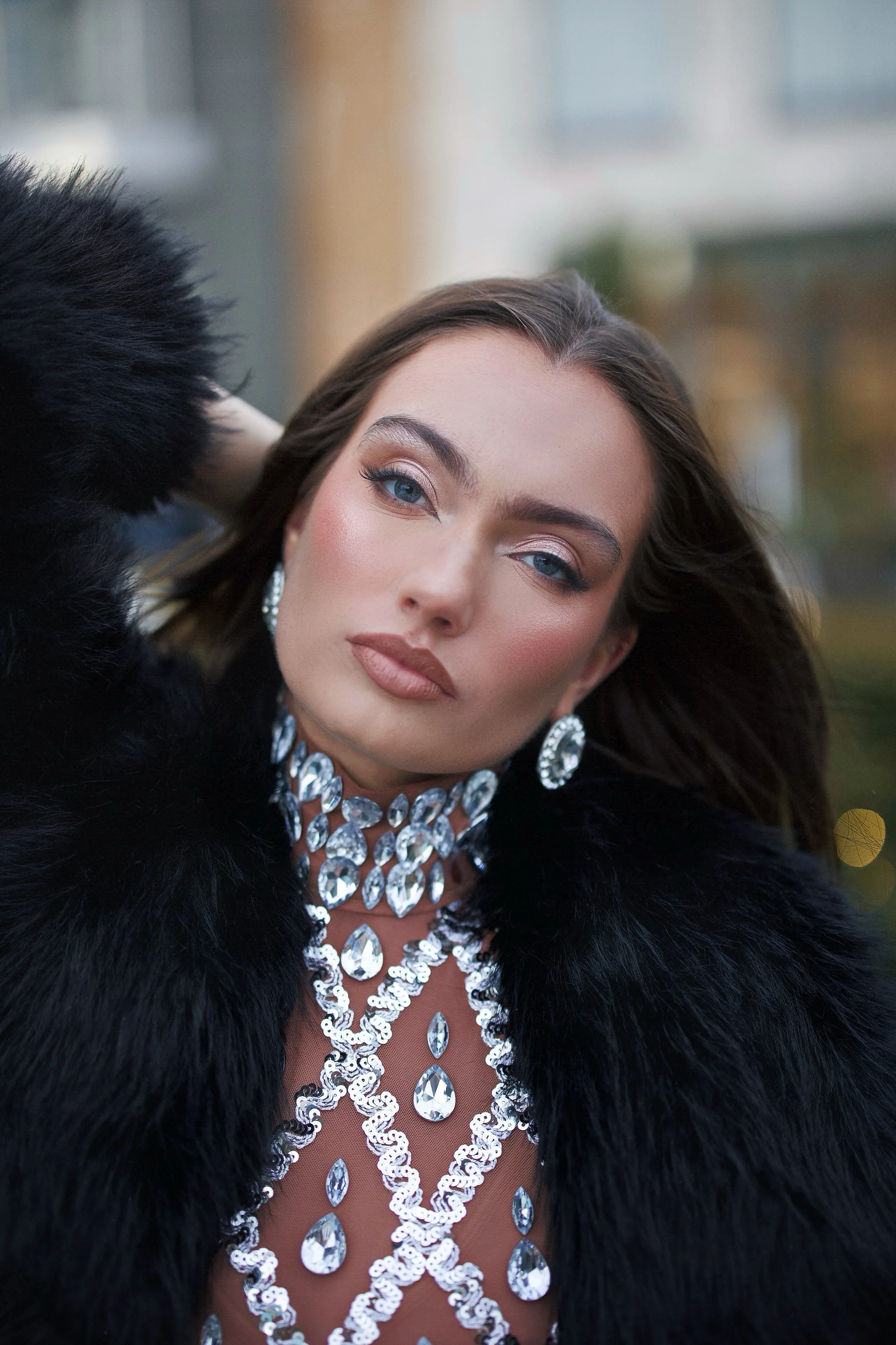 A woman with blue eyes and styled makeup wearing a black fur coat and a sparkling jewelry set with earrings, a necklace, and a gown adorned with large gemstones.