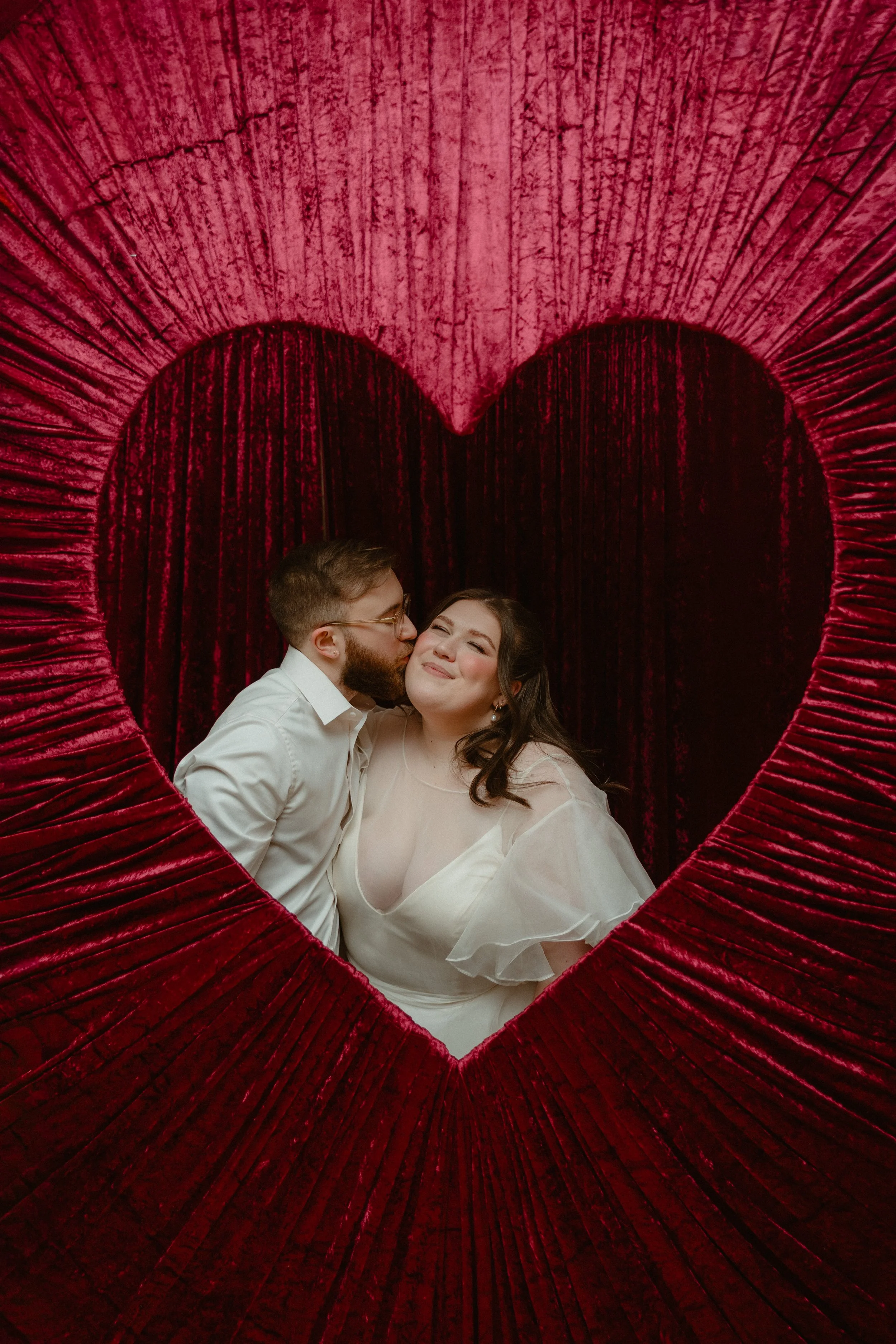 A couple sharing a kiss and smiling, framed by a large red heart-shaped decoration with velvet texture and a dark red curtain background.