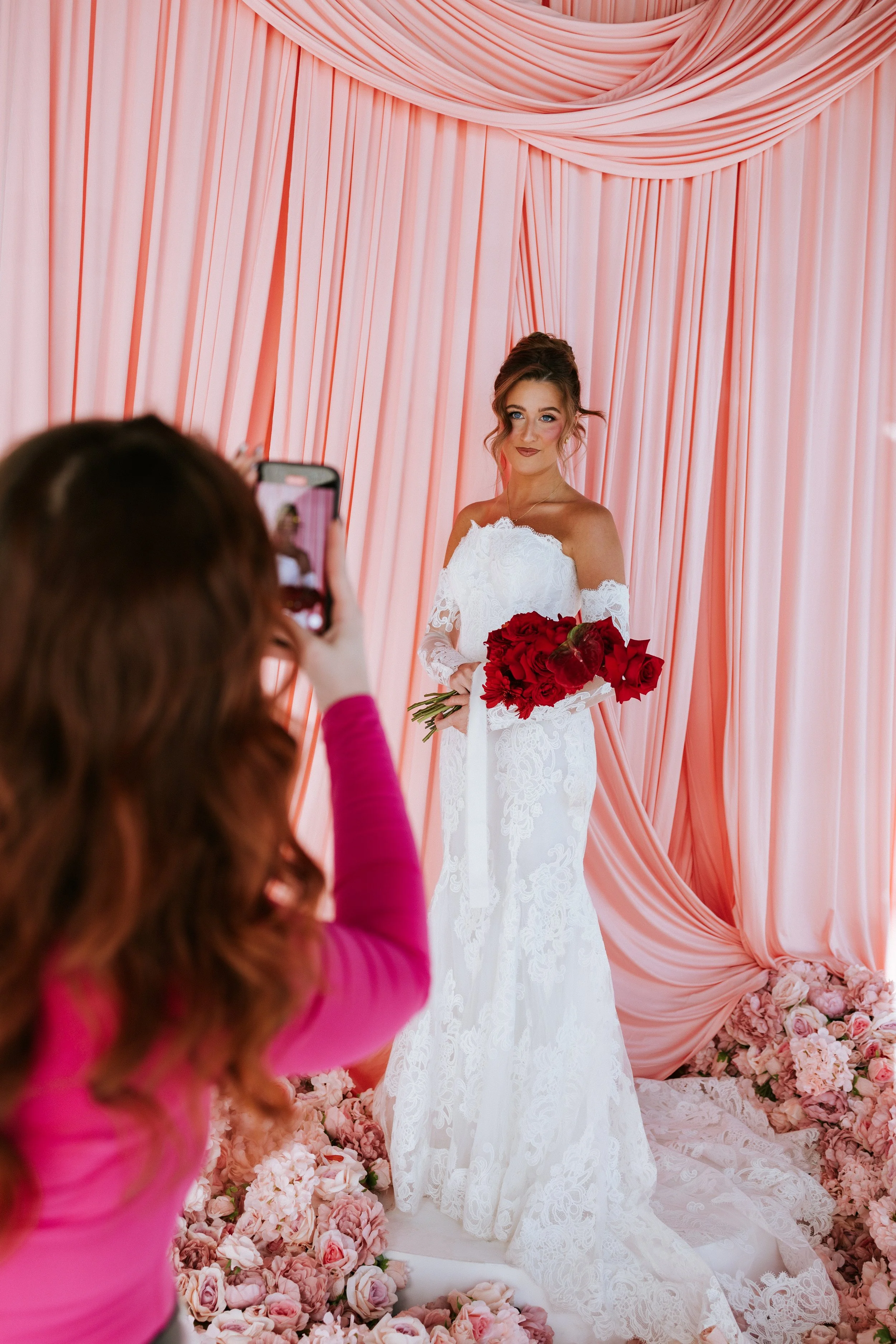 A bride in a white wedding dress holding a bouquet of red roses, standing in front of a pink draped backdrop with floral decorations at her wedding.
