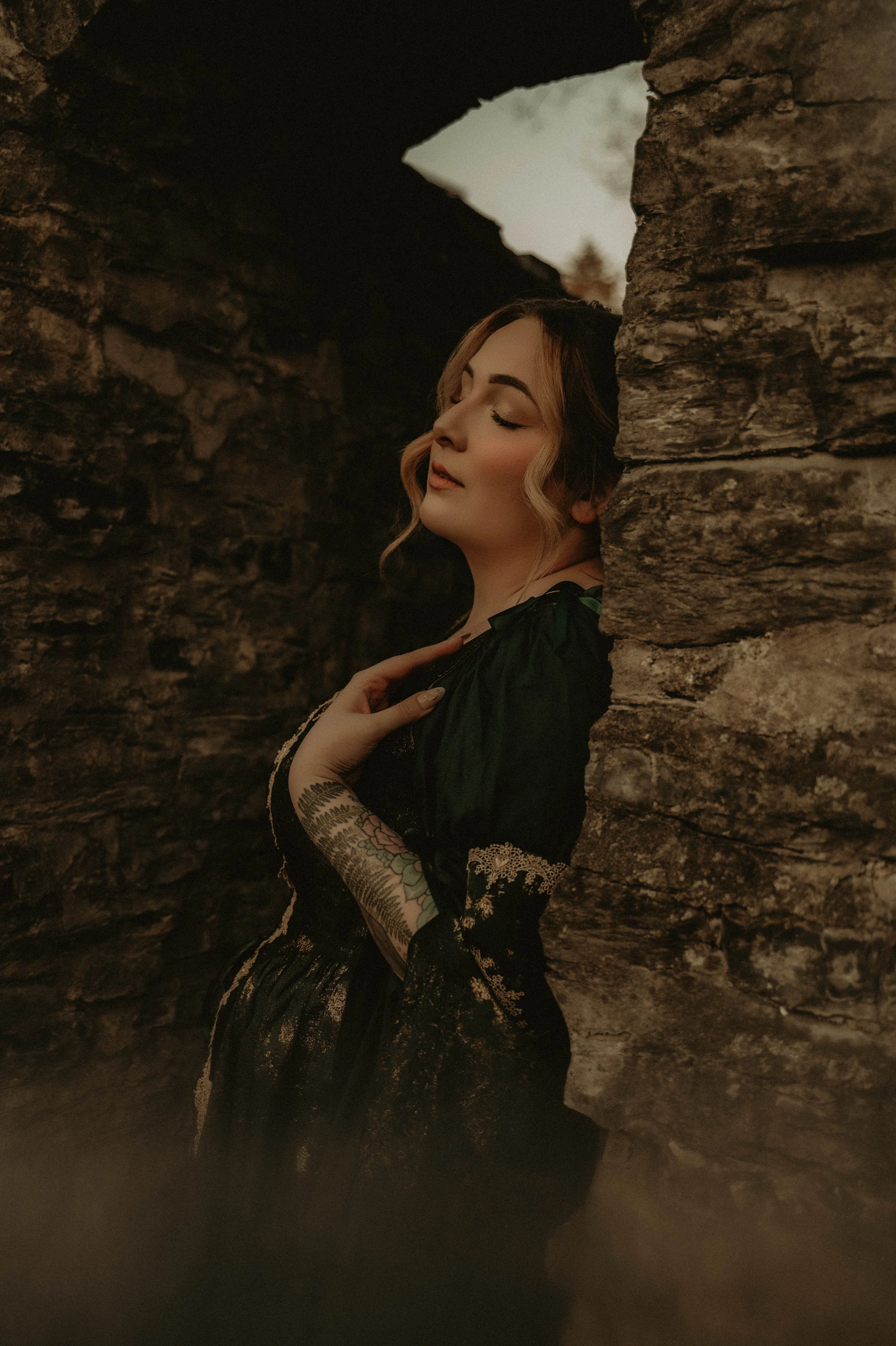 A woman with light skin and gemstone makeup, wearing lace and embroidery clothing, is posed between stone walls with her eyes closed.