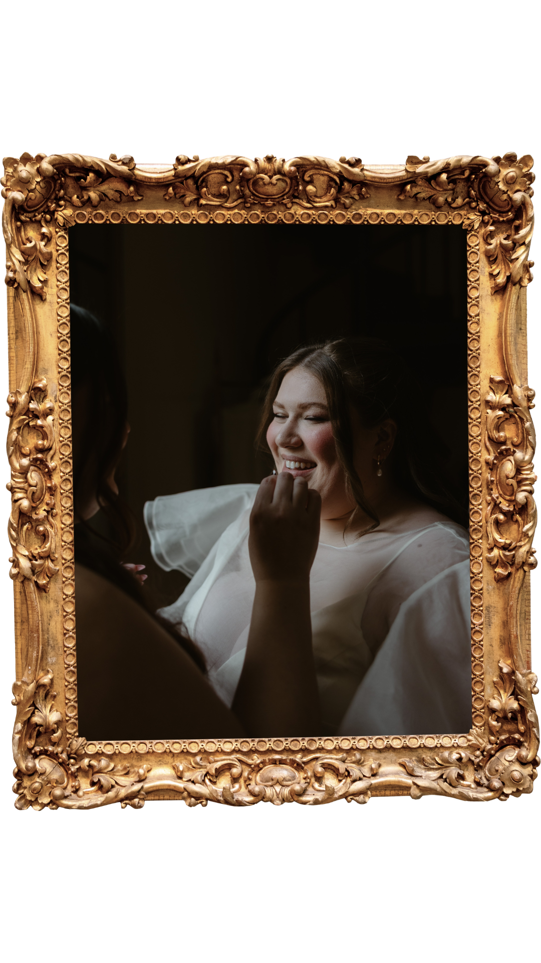 A woman in a white dress smiling as another person applies makeup or lipstick to her lips, reflected in an ornate gold framed mirror.