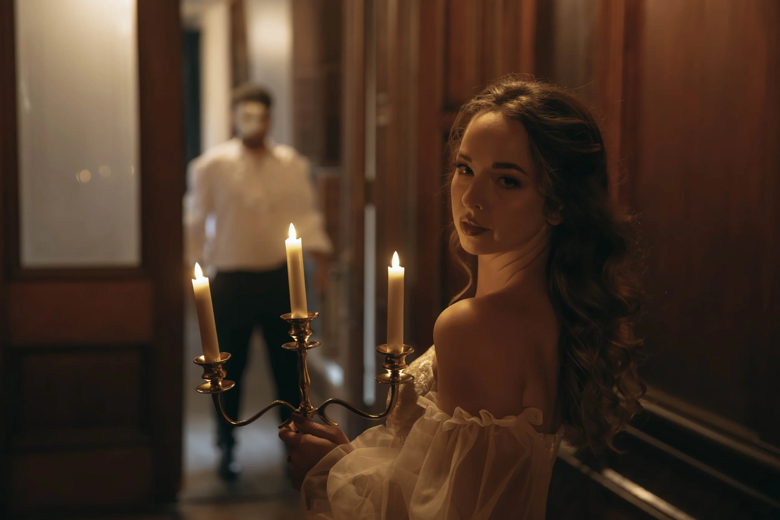 A woman with long, curly hair holding a gold candelabrum with three lit candles in a dimly lit room.